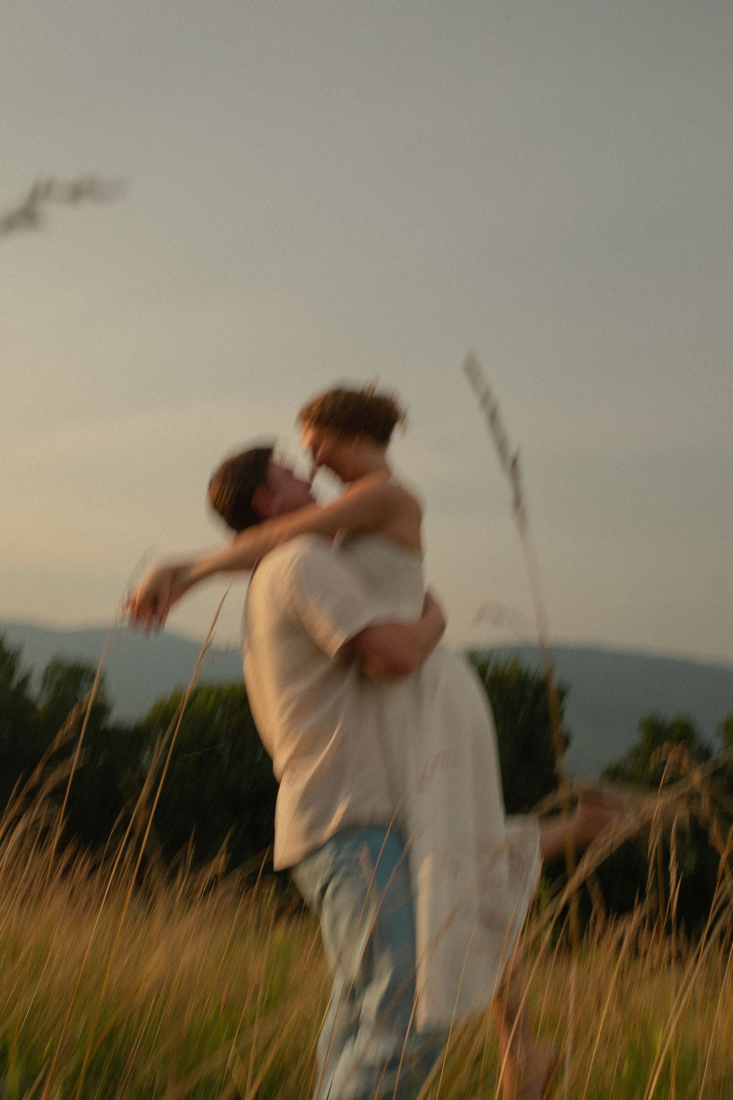 Couple embracing romantically in golden meadow at sunset, motion blur effect, mountains in background.
