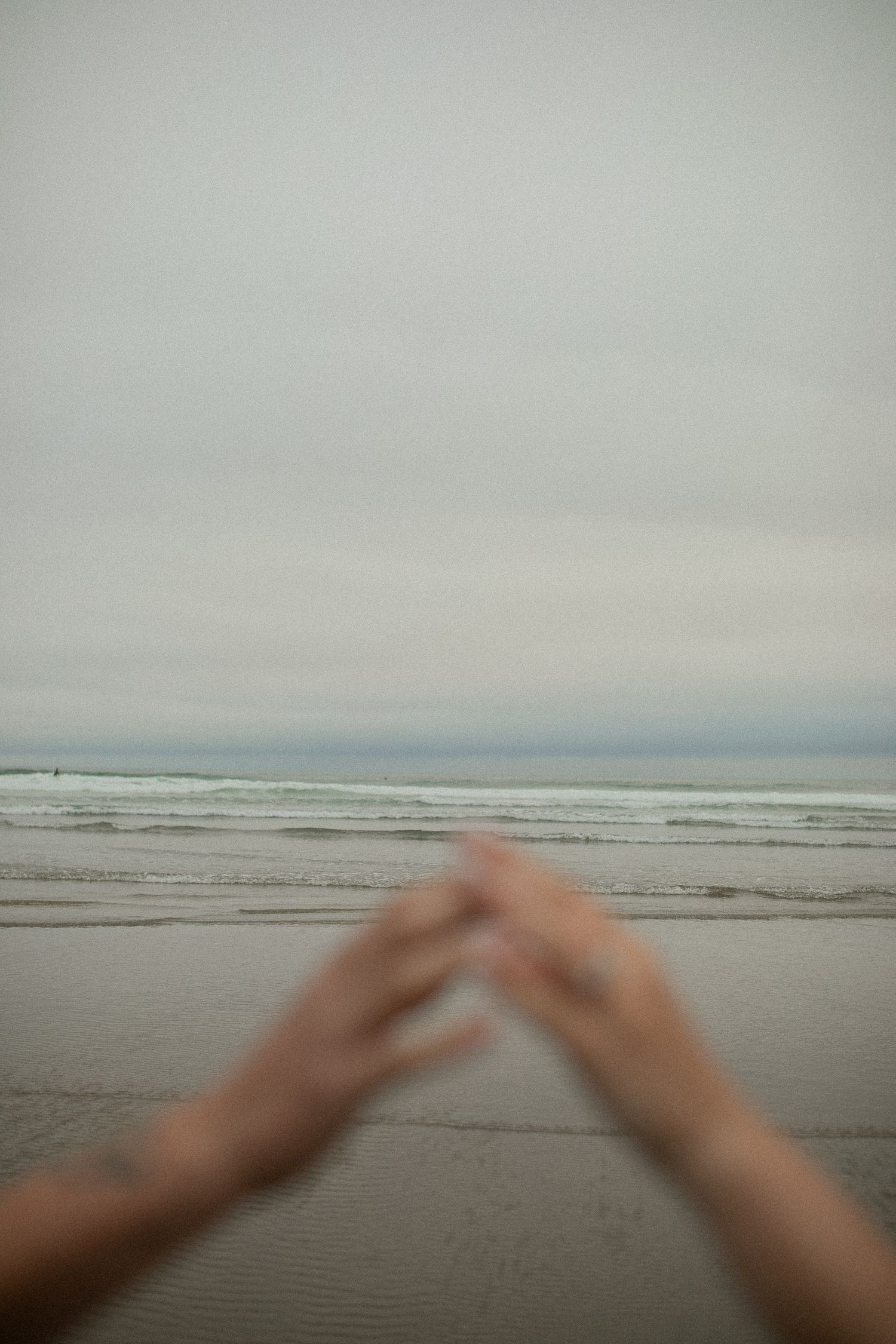 Blurred hands reaching toward a misty ocean beach with overcast skies and gentle waves in the background.