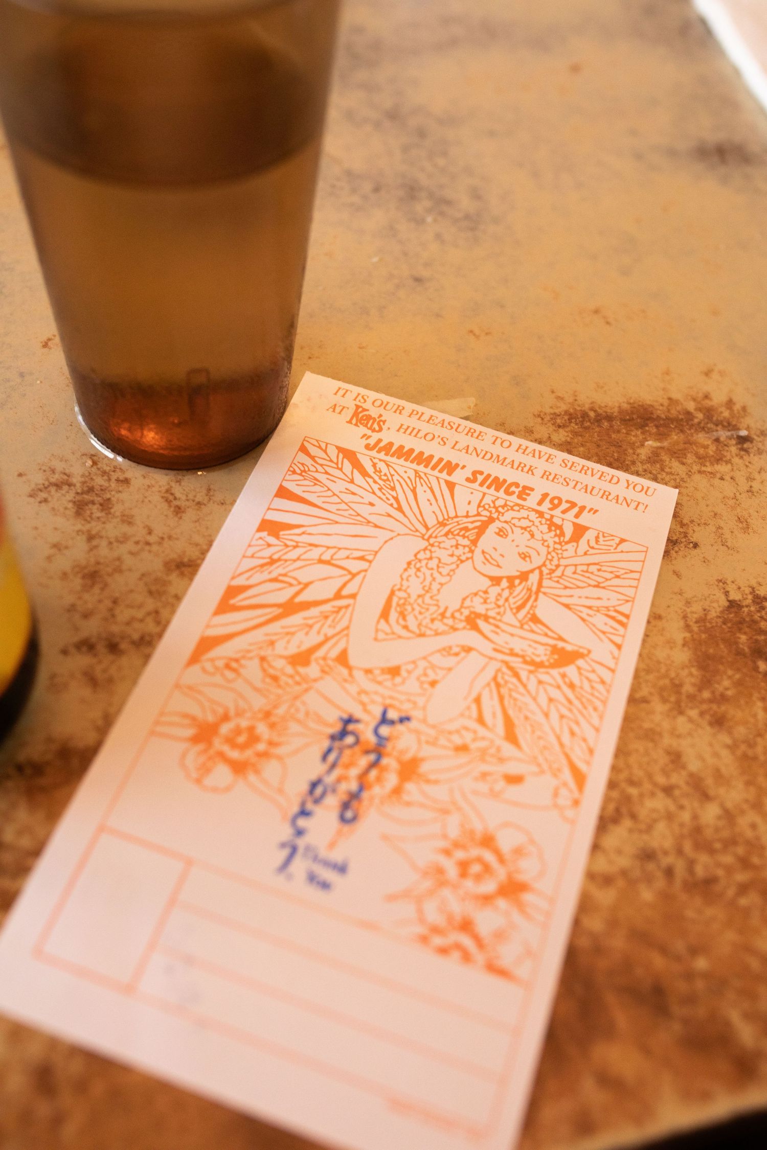 A beer glass and an orange-tinted receipt or ticket with decorative floral design sit on a weathered surface.