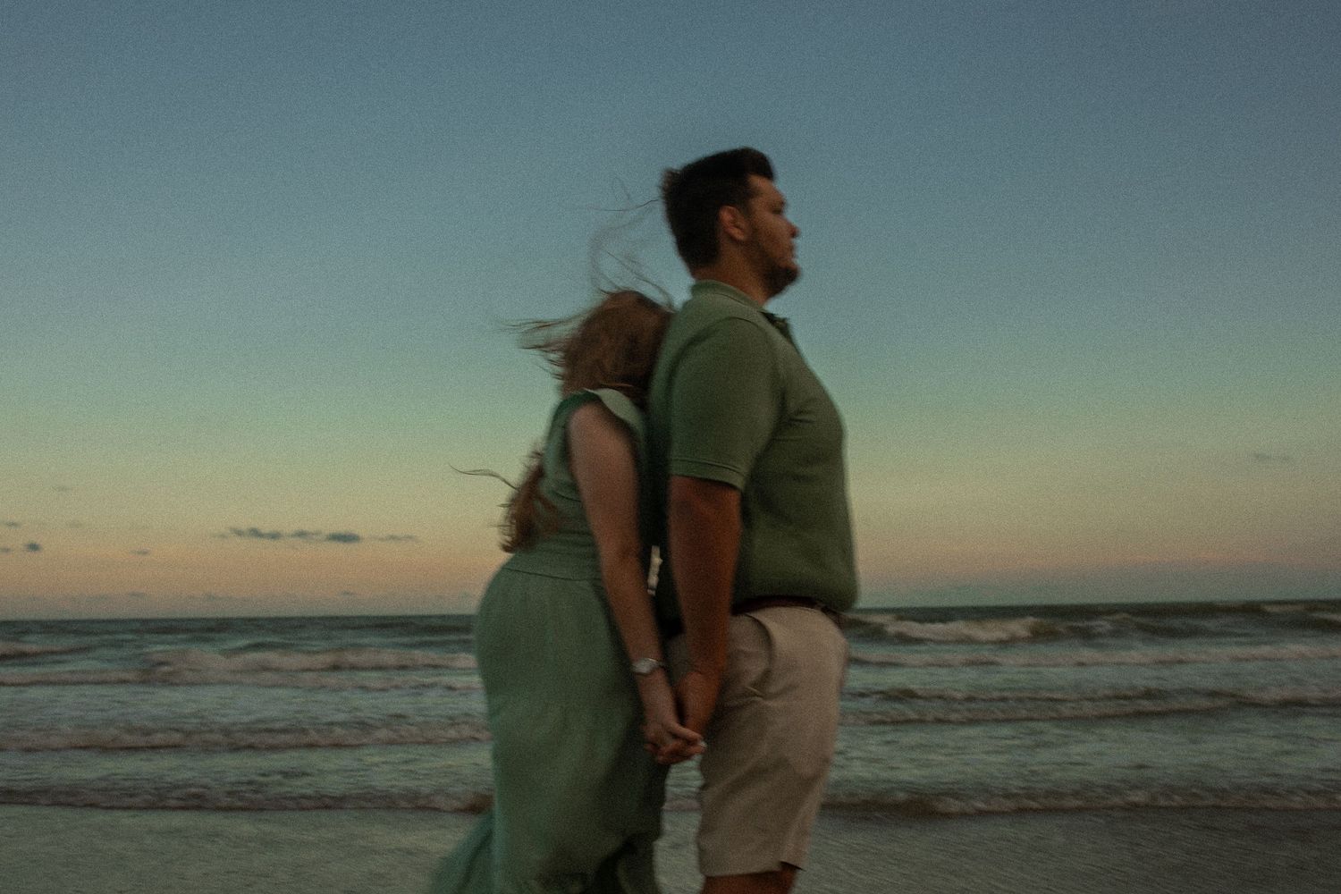 A romantic couple stands back to back on a sandy beach during sunset as waves roll in on the shore.
