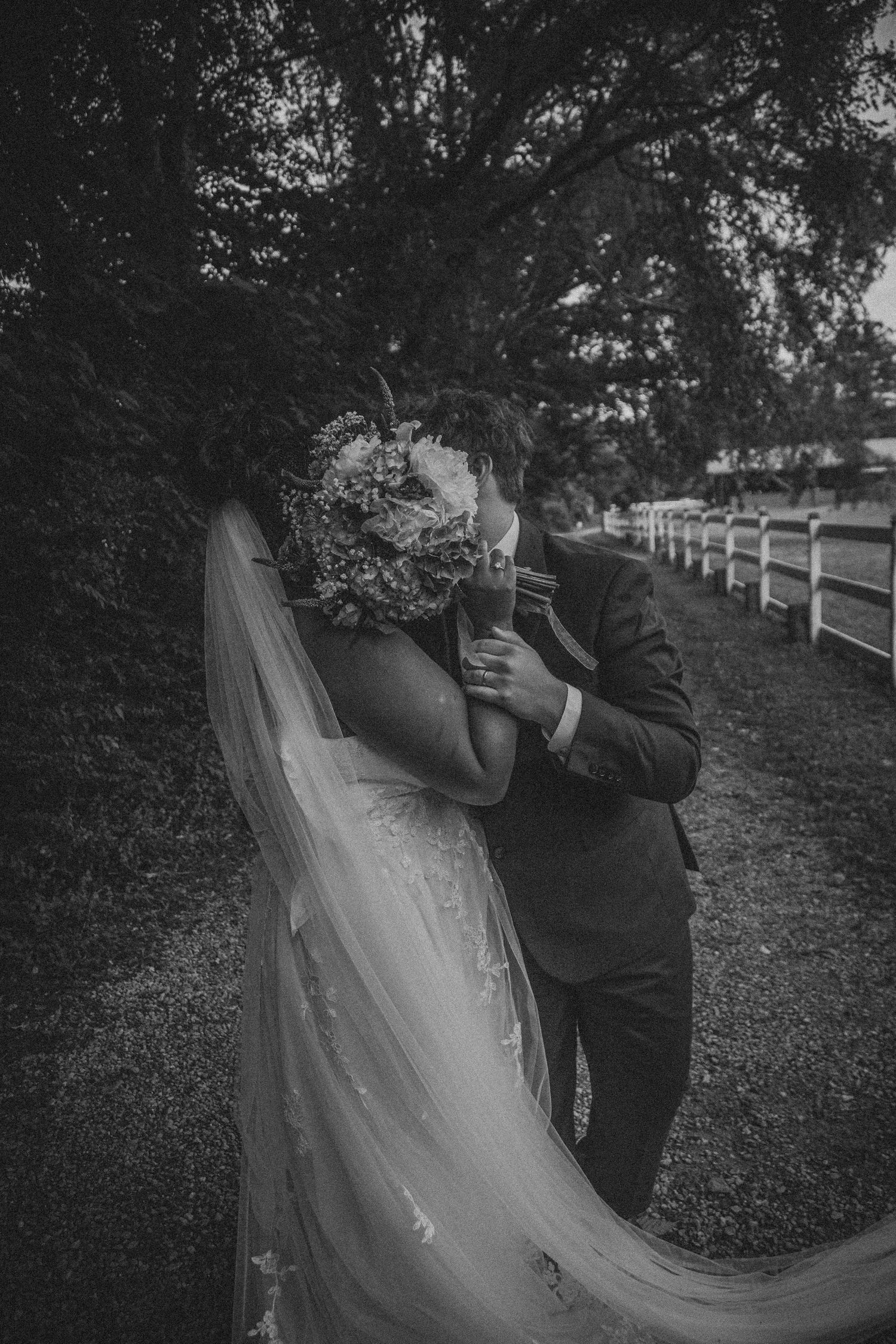 Black and white photo of a bride and groom kissing behind a floral bouquet on a scenic path.