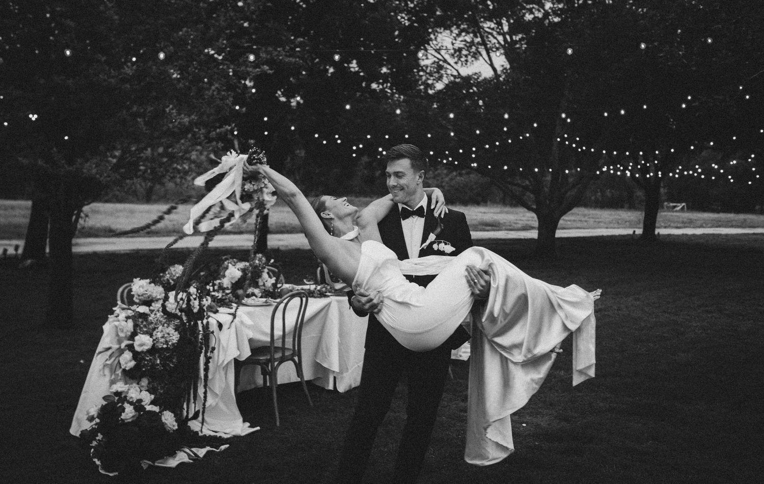 Black and white wedding photo of groom carrying bride in elegant gown outdoors under string lights.