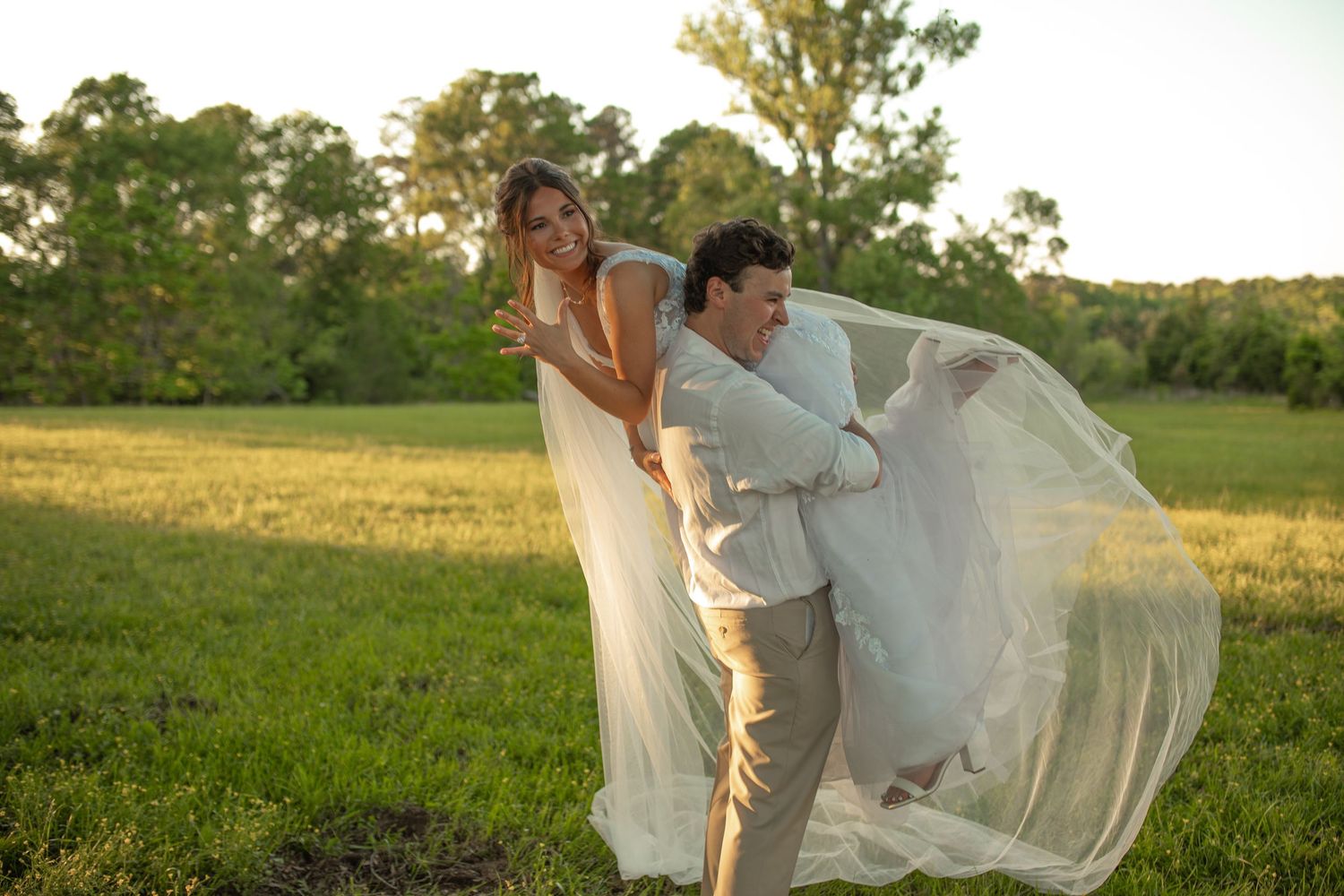 A groom playfully lifts his bride in a white flowing gown outdoors in a lush green field at golden hour.