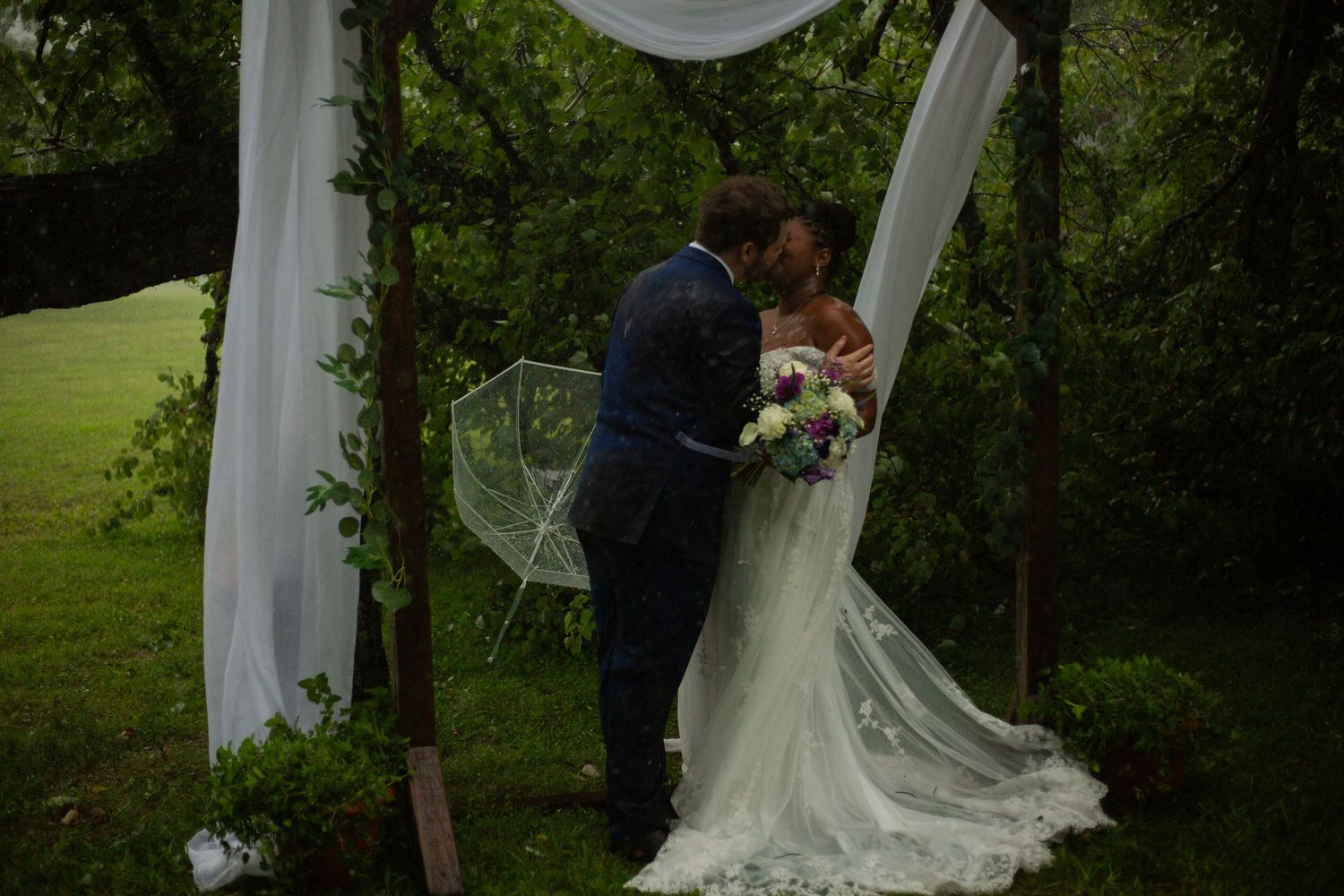 Bride and groom sharing a kiss under a rustic wooden arch with white draping during an outdoor garden wedding ceremony.
