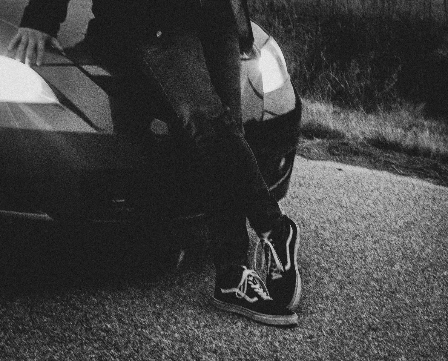 A black and white close-up of shoes and legs next to a car on a road.