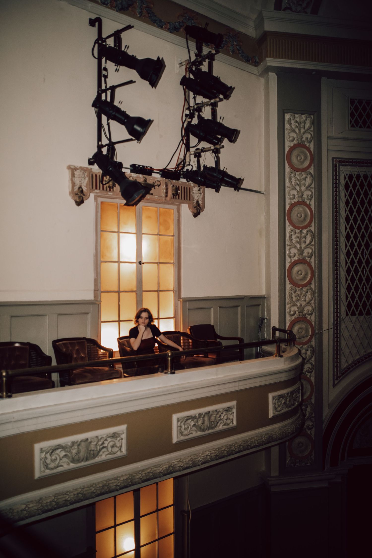 Dimly lit theater balcony with vintage lighting fixtures casting warm glow through windows and ornate architectural details.