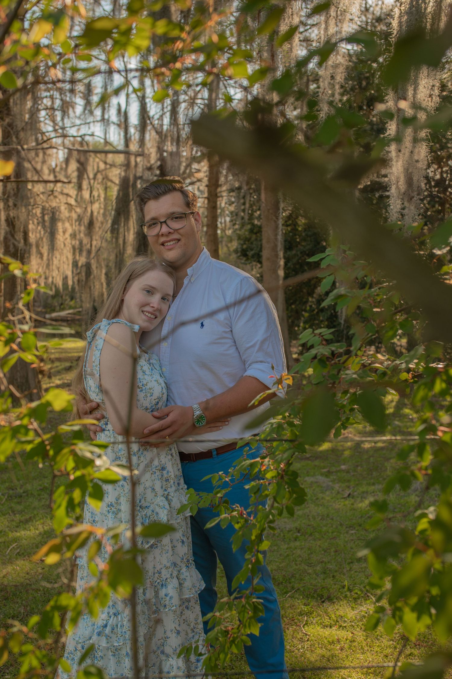 A romantic couple embraces in a natural setting framed by fall foliage and sunlit trees in the background.