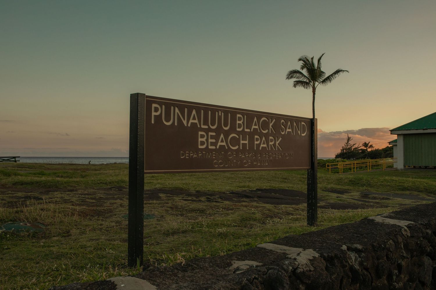 A wooden sign for Punaluu Black Sand Beach Park stands against a sunset sky with a palm tree silhouette in Hawaii.