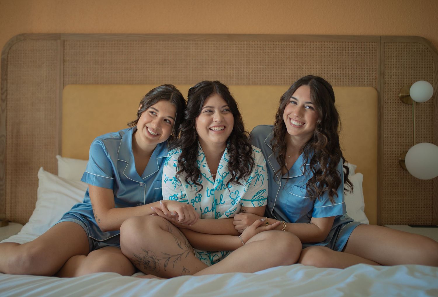 Three smiling women in matching blue pajamas sitting together on a white bed in a bright hotel room.