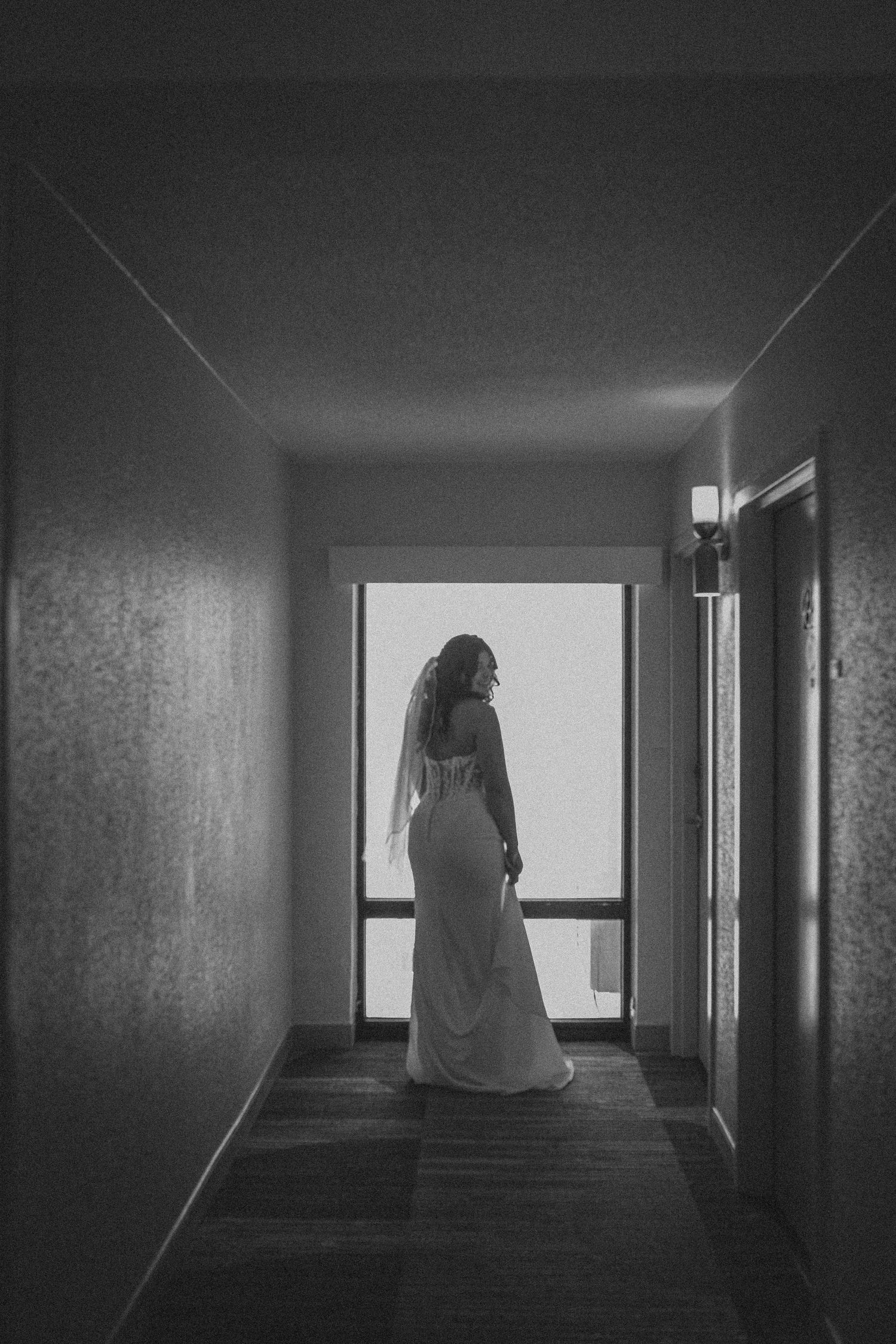 Black and white photo of a bride in a white gown and veil standing in a dimly lit hallway near a bright window.
