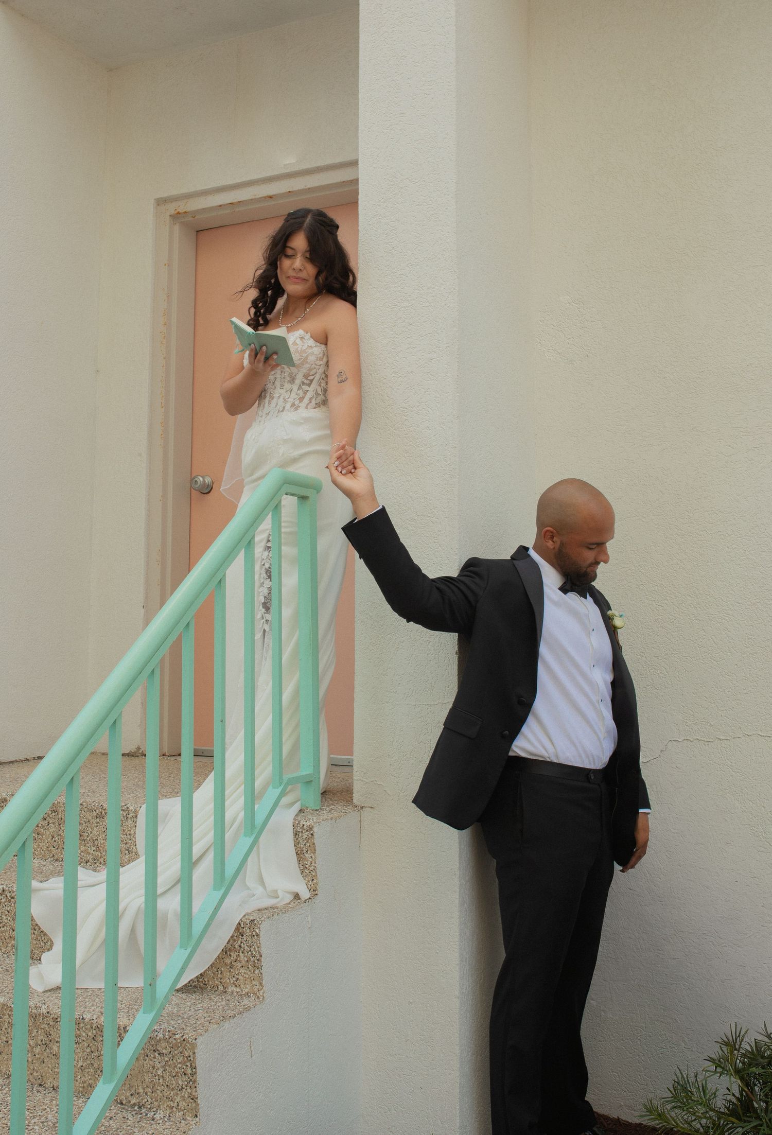 Bride in white gown reads vows on stairs while groom in tuxedo holds her hand around a corner before their first look.