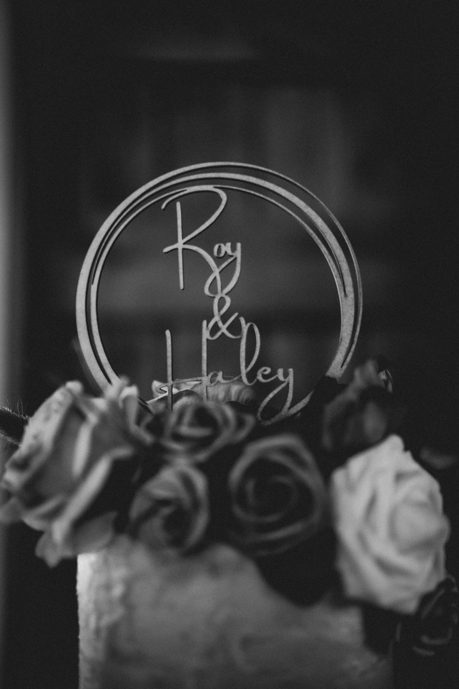 Black and white wedding detail photo showing a circular name sign surrounded by roses on a cake topper.