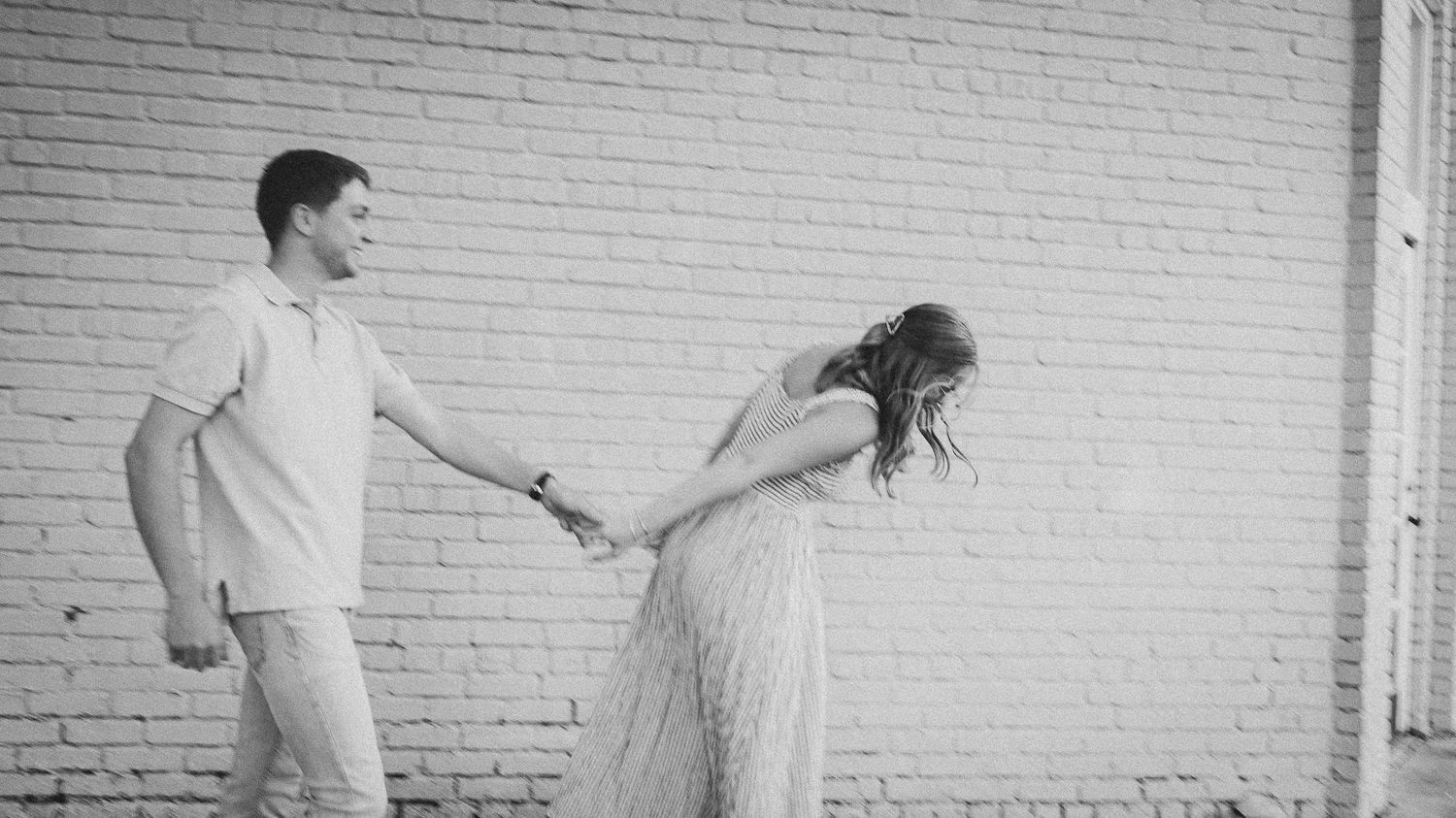 A couple in light-colored outfits holds hands while leaning away from each other against a white brick wall.