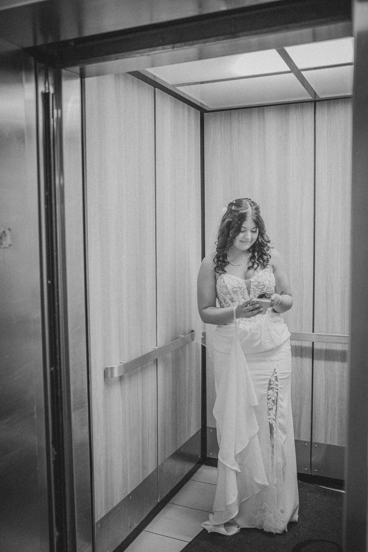 Black and white photo of a bride in a wedding dress checking her phone inside an elevator.