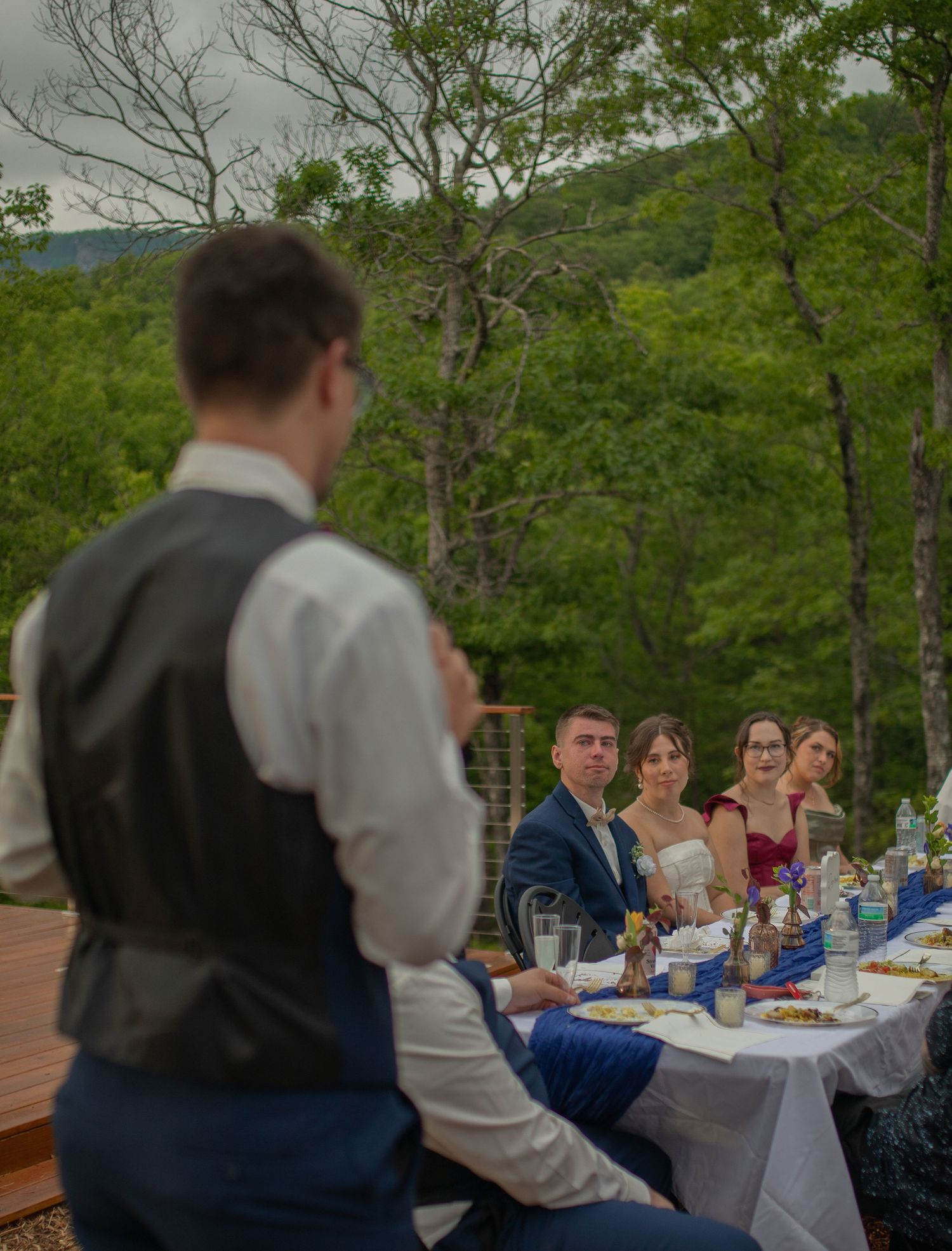 Groom giving a toast speech at an outdoor wedding reception dinner table surrounded by lush green trees.