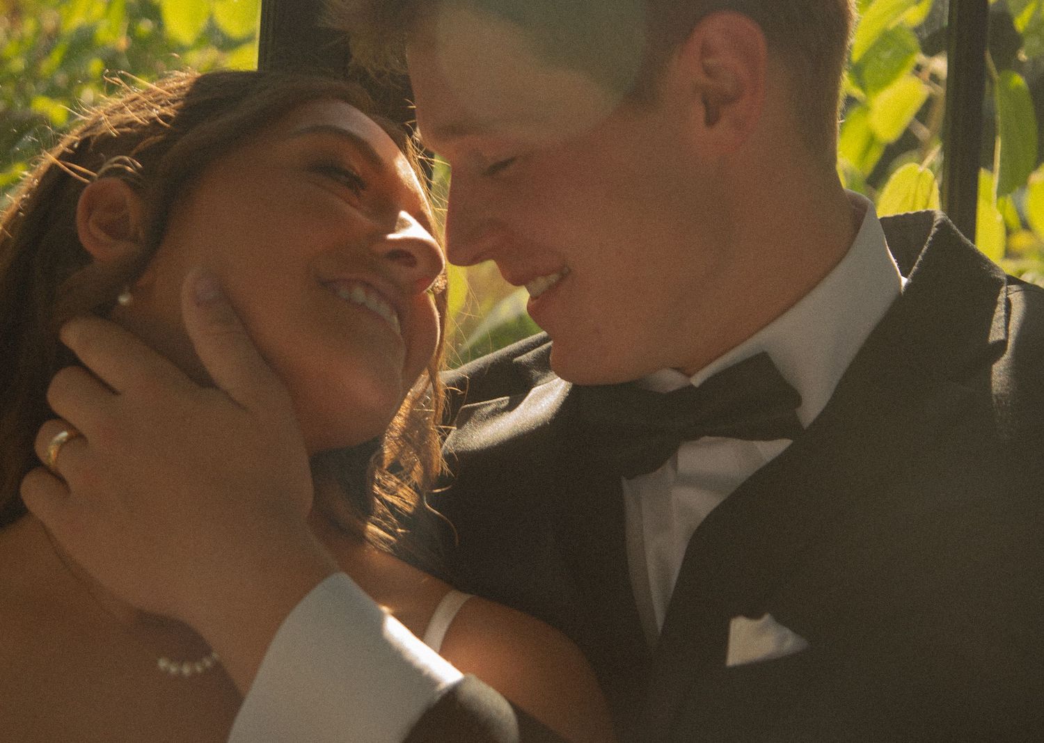 A bride and groom share a romantic moment, smiling closely together in soft golden sunlight outdoors.