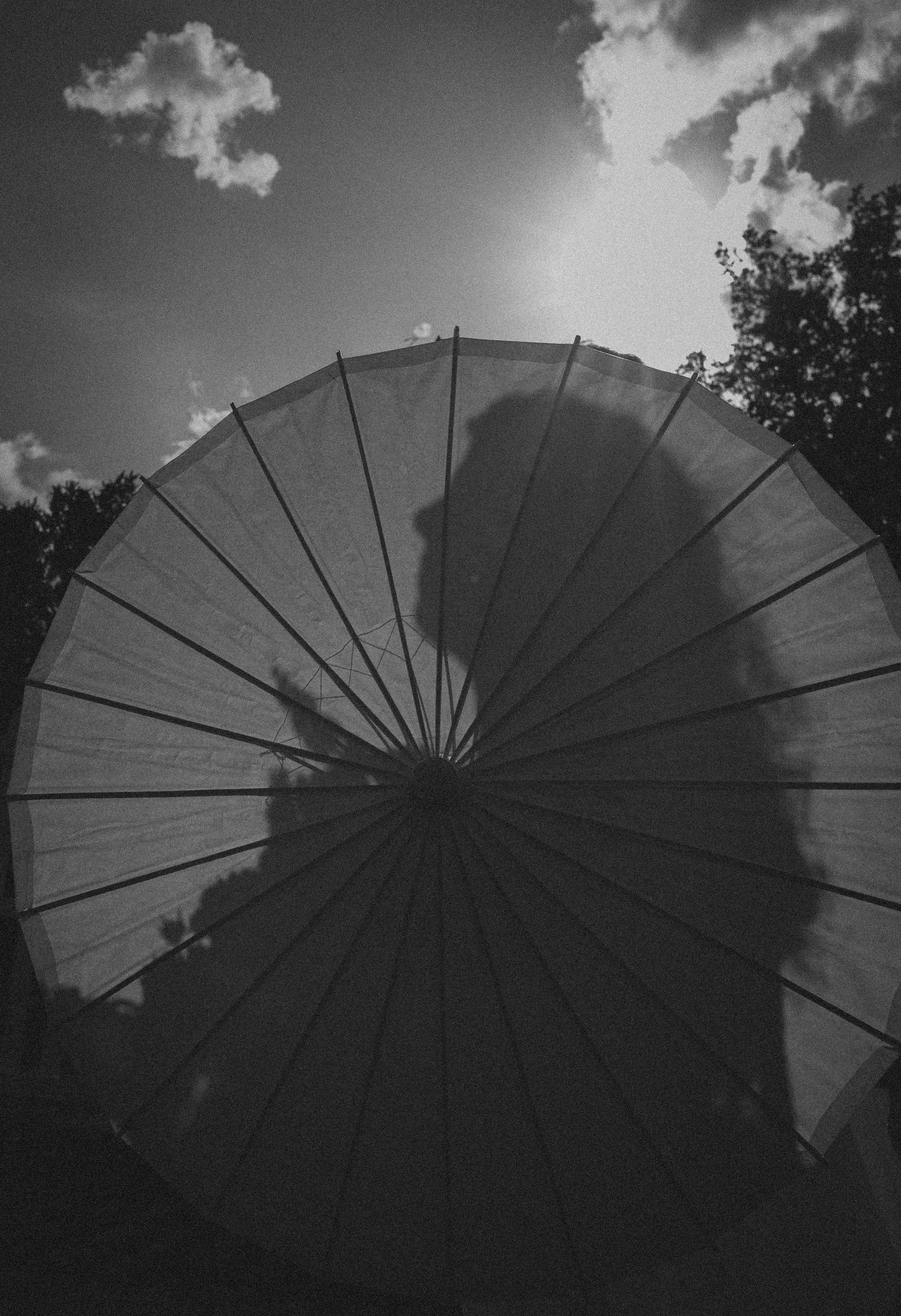 Black and white photo of a large parasol umbrella backlit by sun, casting a shadow silhouette against cloudy sky.