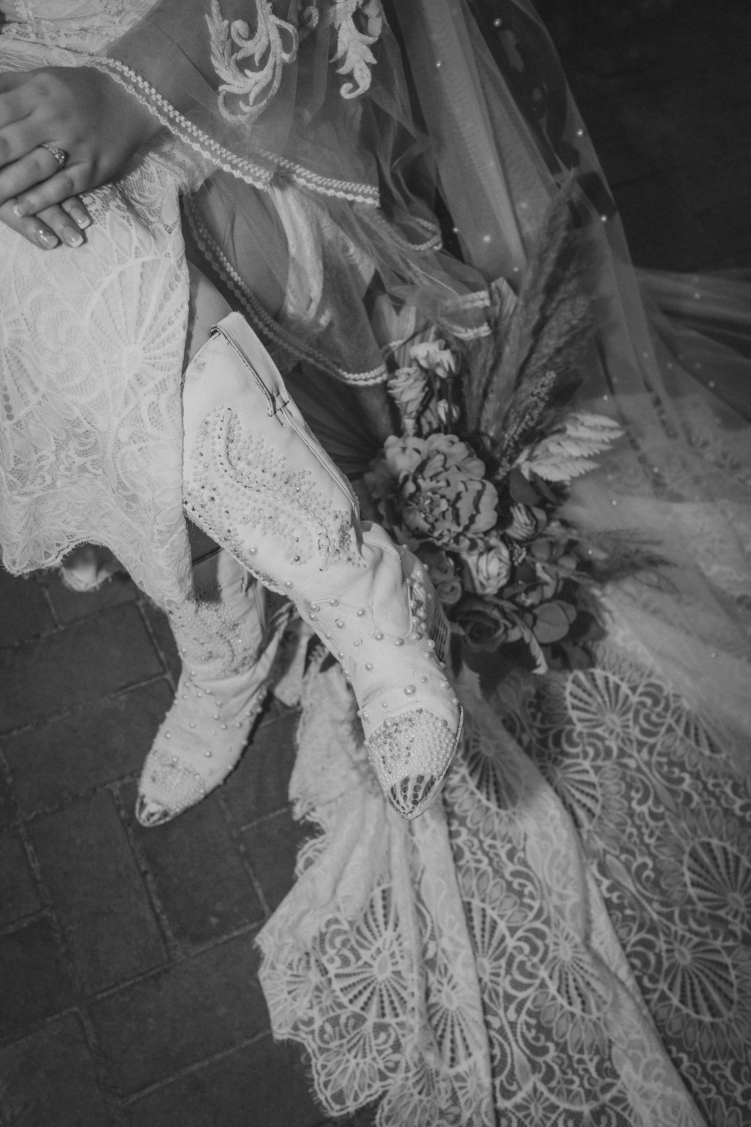 A black and white wedding detail photo showing lace dress, vintage boots and a floral bouquet resting on ornate lace fabric.