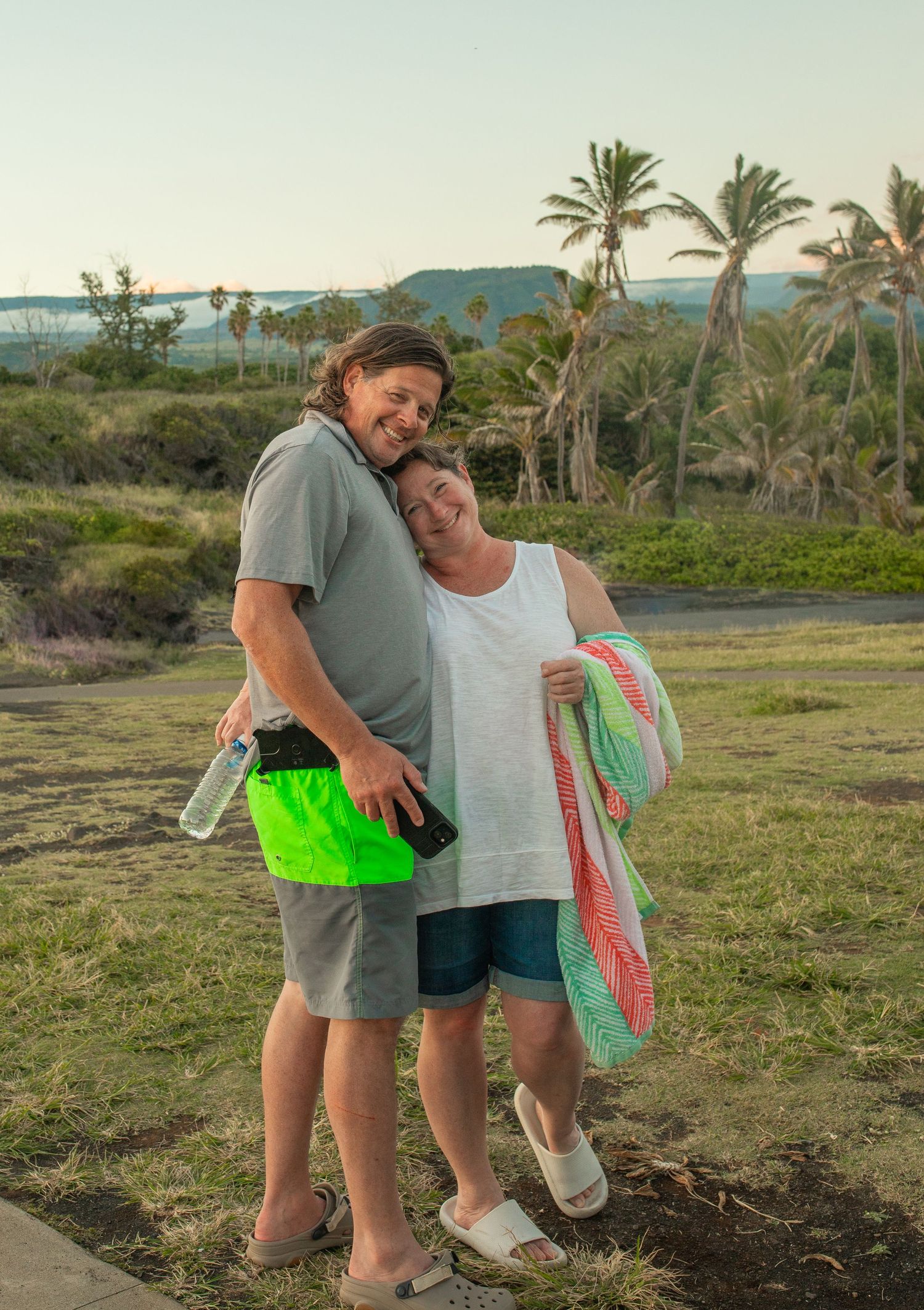 A couple shares an affectionate moment on a scenic path with palm trees and mountains in the background at sunset.