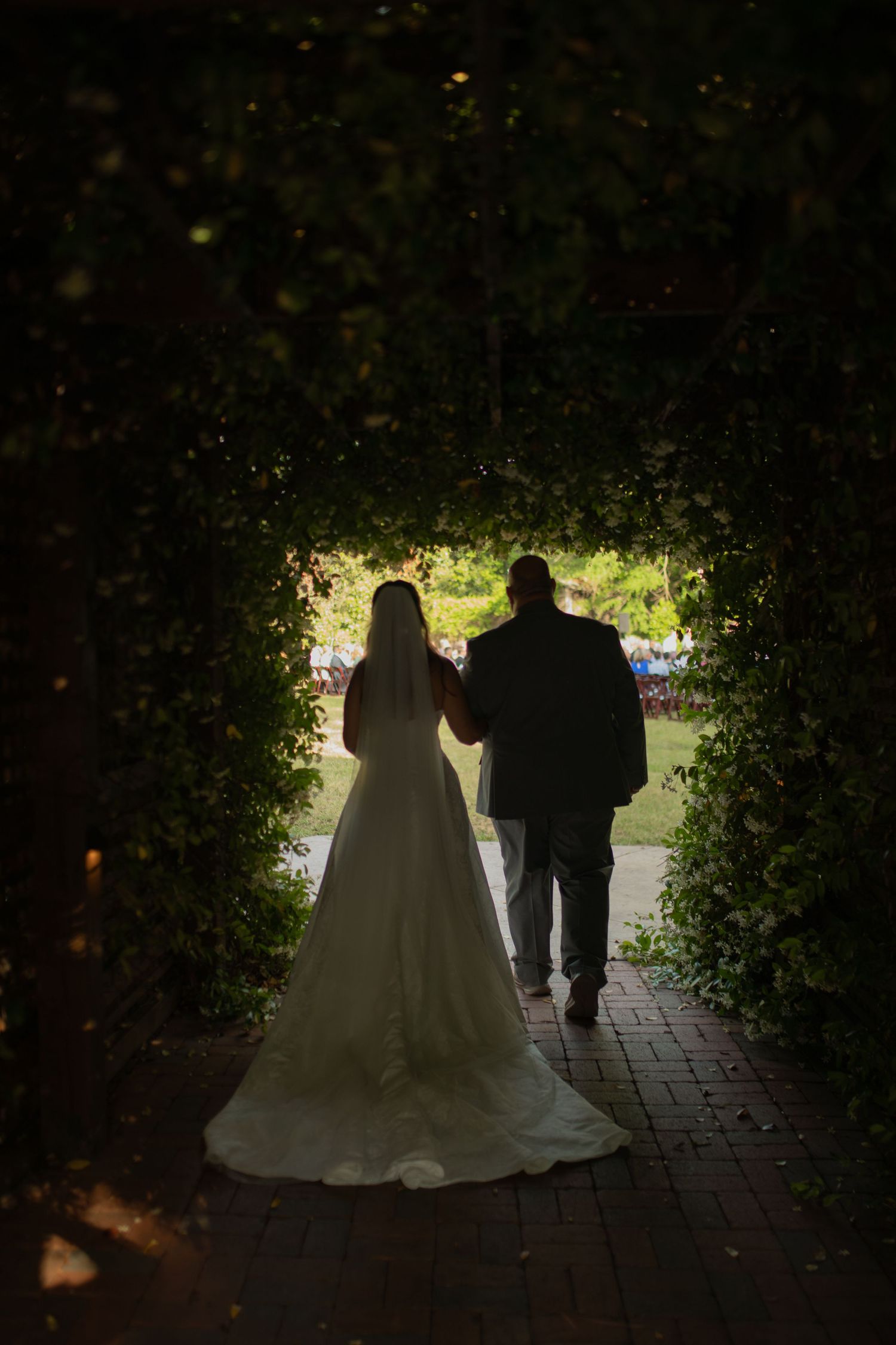 Bride and groom silhouetted walking through a lush green garden archway on their wedding day.