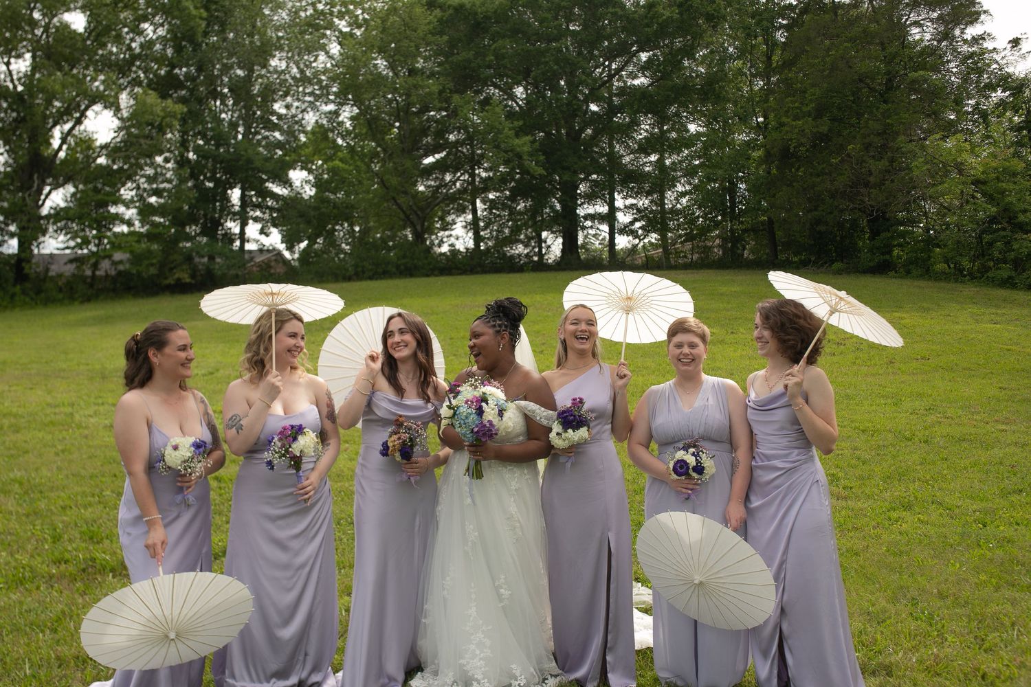 Bride in white gown with six bridesmaids in lavender dresses holding white parasols on a green lawn.