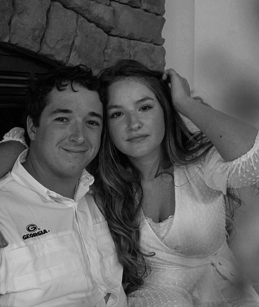 Black and white photo of a young couple smiling together near a stone fireplace, man wearing a Georgia shirt.