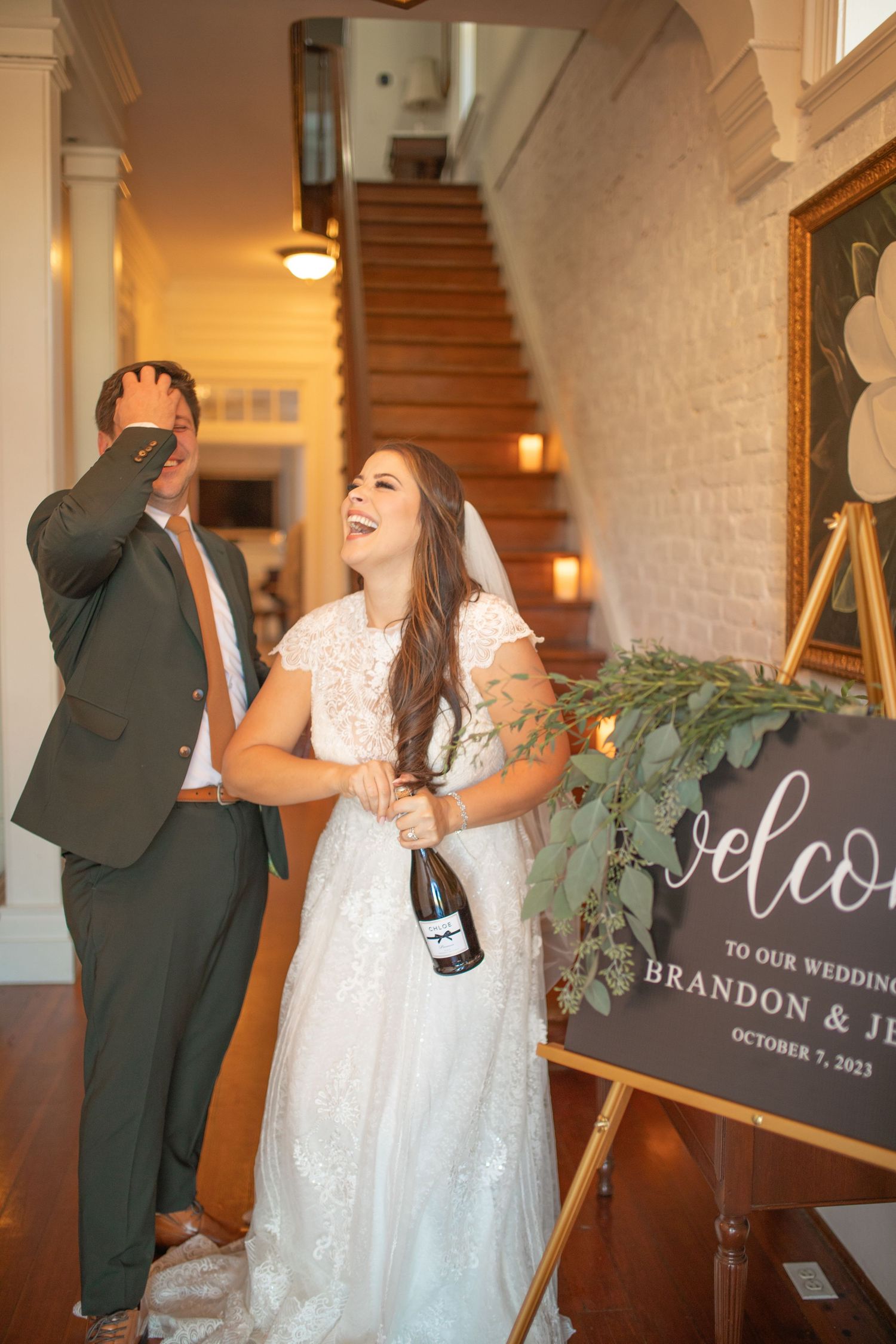 Bride and groom laughing joyfully at wedding welcome sign reading Brandon and Je, October 7, 2023, holding champagne bottle.