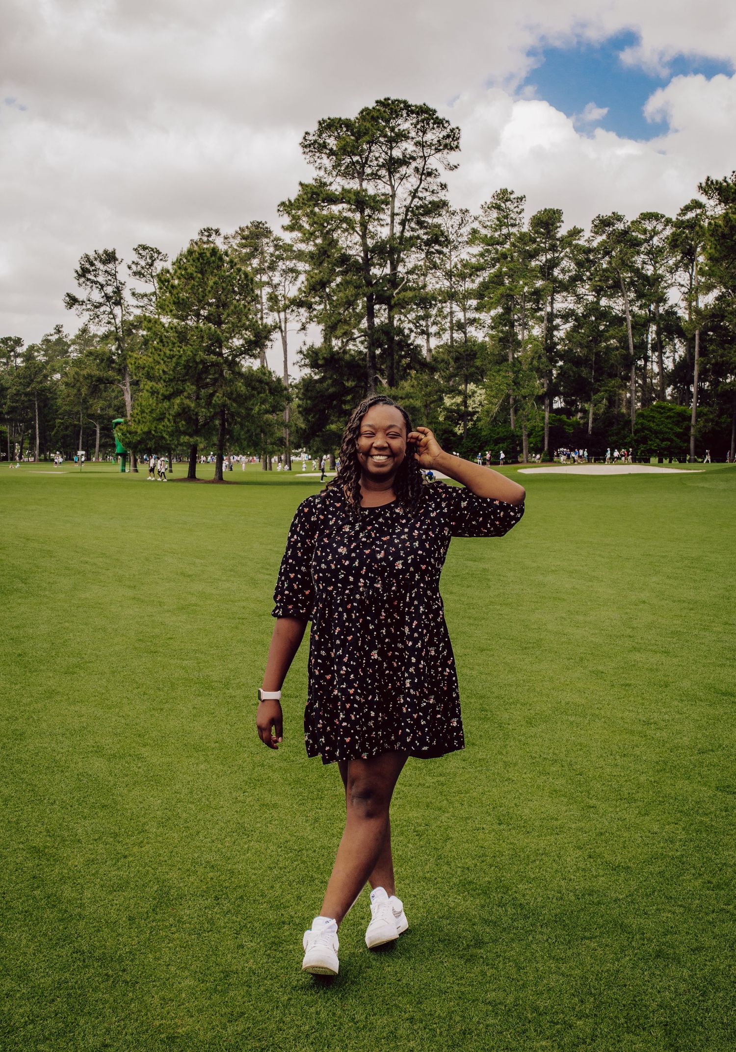 Someone wearing a polka dot dress and white sneakers poses on a lush green lawn with tall pine trees in the background.