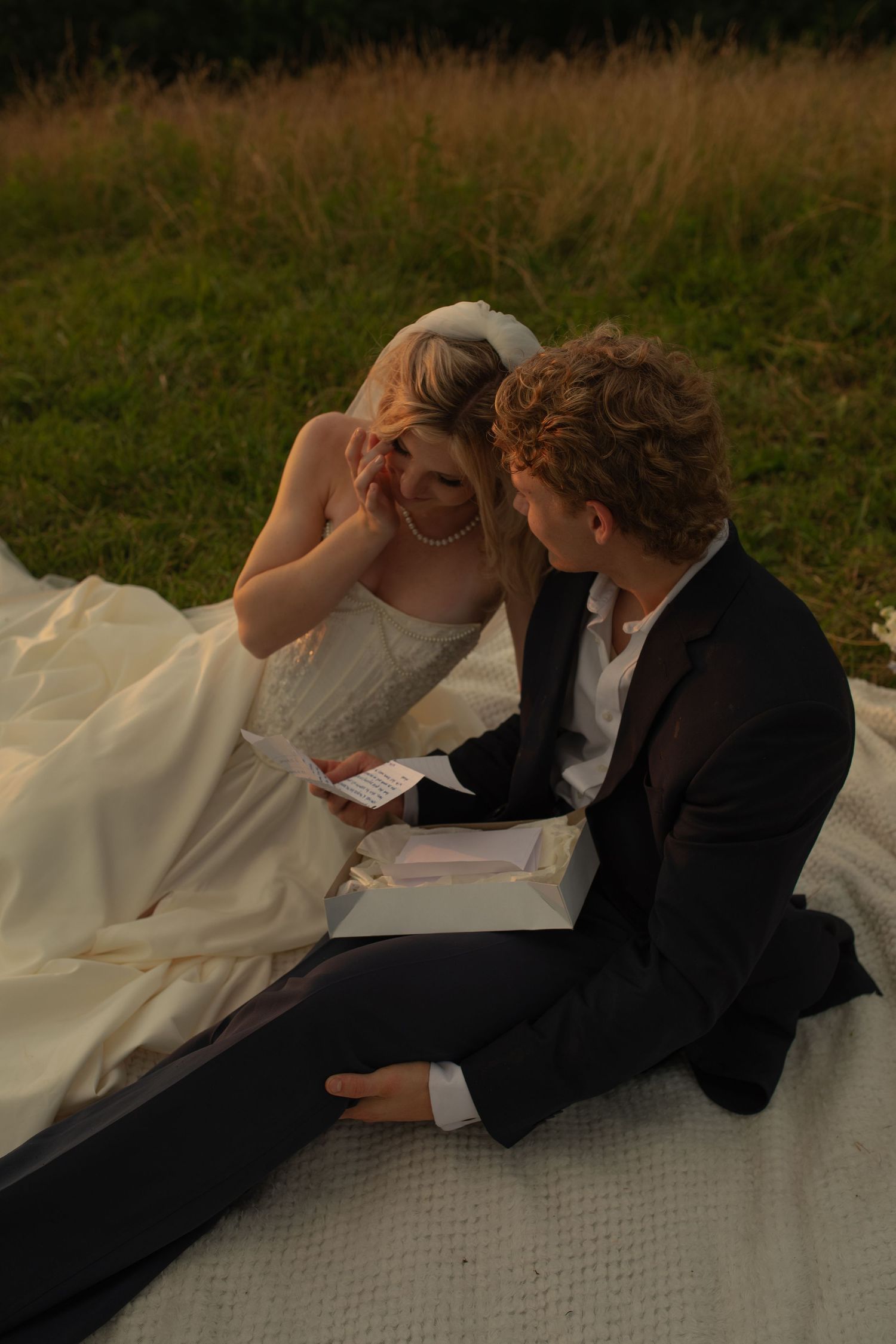 Bride and groom sitting on white blanket in field, sharing intimate moment with open gift box at golden hour.