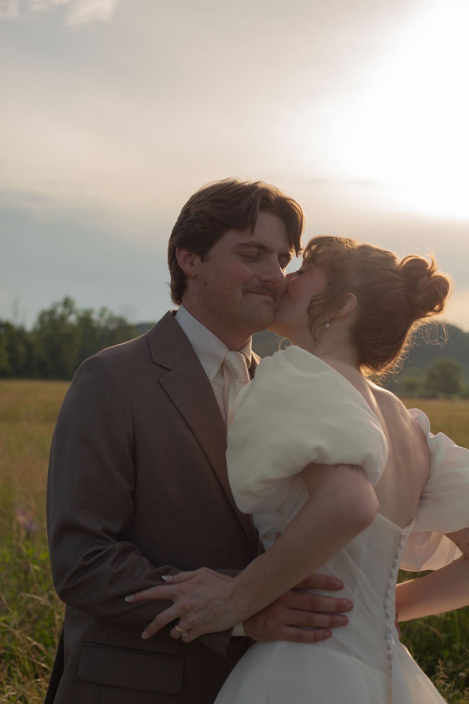 A bride in white kisses her groom in a gray suit during a romantic outdoor wedding at golden sunset.