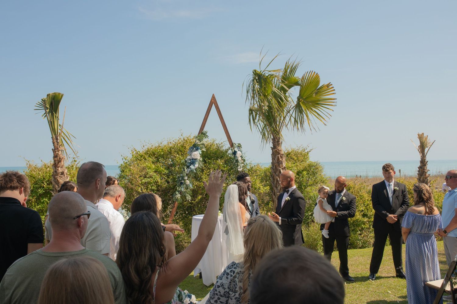 Outdoor beach wedding ceremony with couple at triangular arch, guests seated on lawn, palm trees and ocean in background.