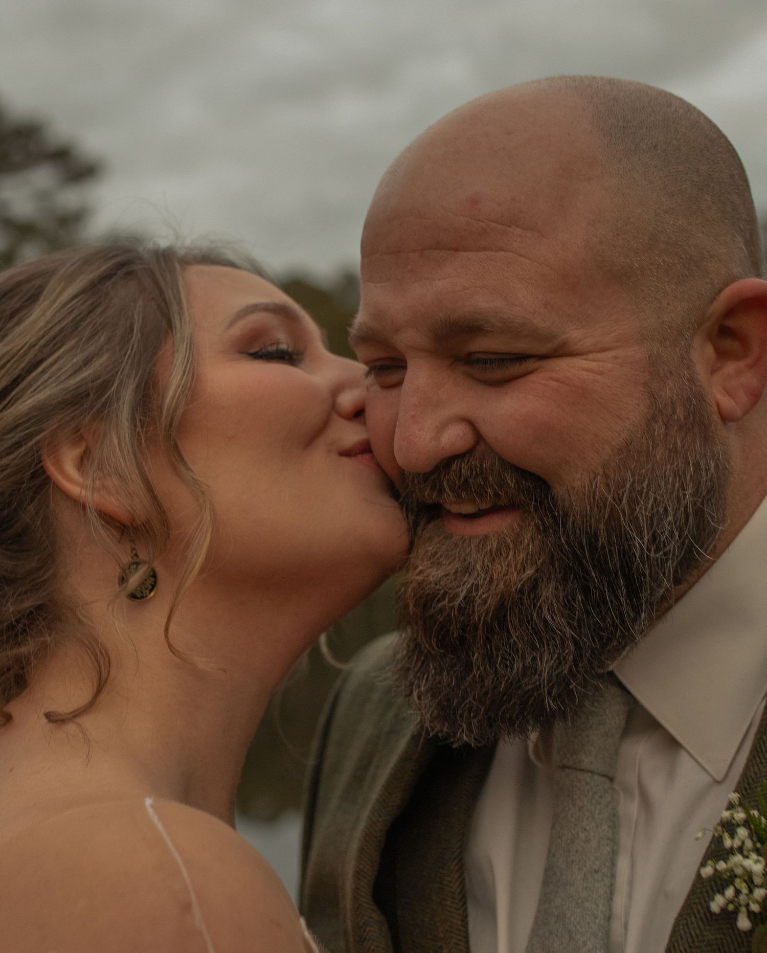 A bride kisses the cheek of her smiling bearded groom on their wedding day outdoors.