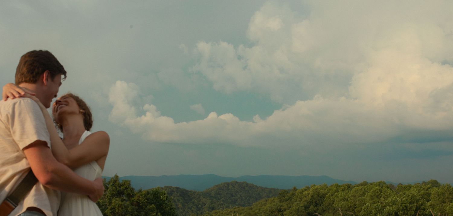 A couple embraces on a scenic overlook with lush green mountains and dramatic cloudy sky in background.
