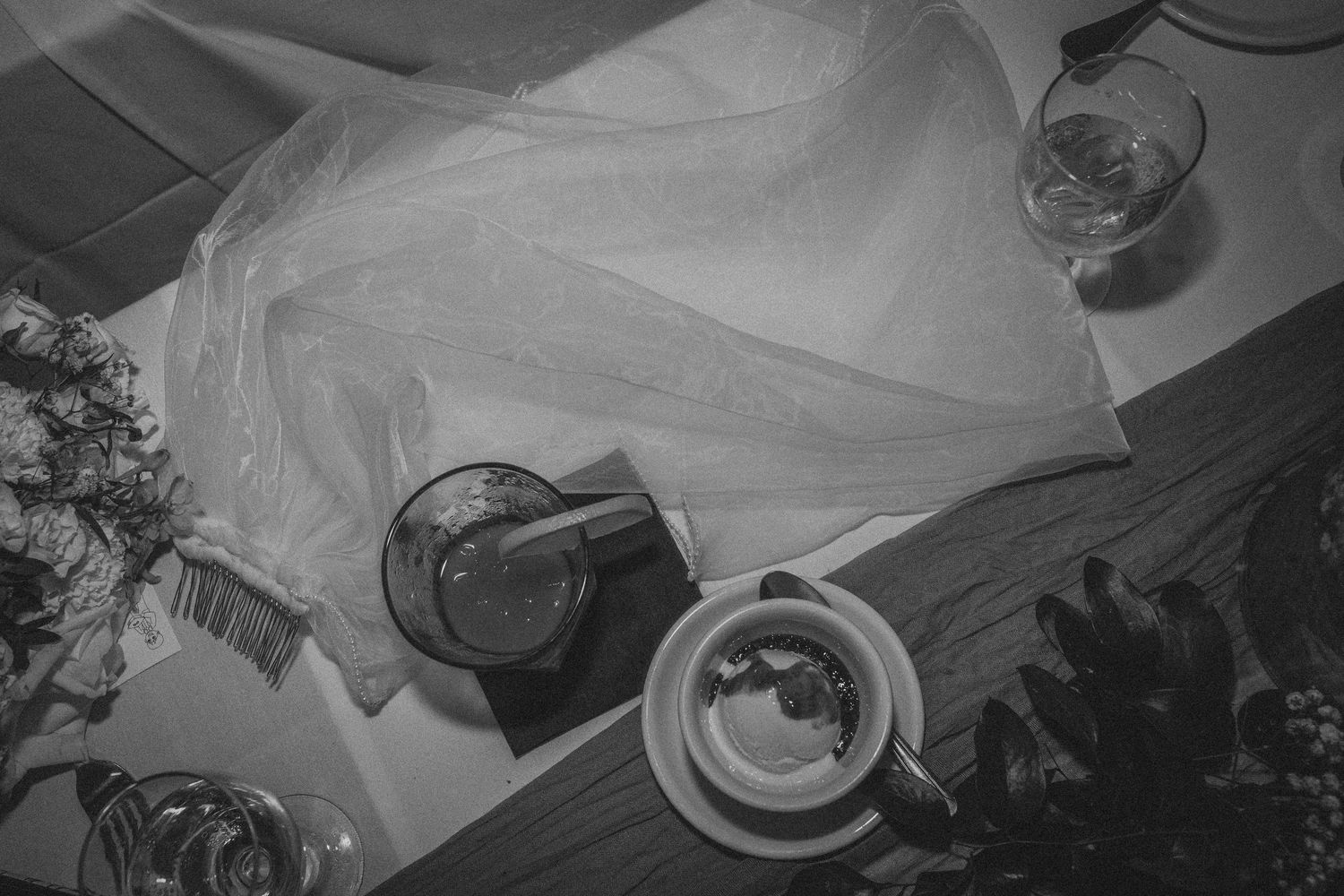Black and white overhead view of a table setting with tea cup, drinks, and white napkin on wooden surface.