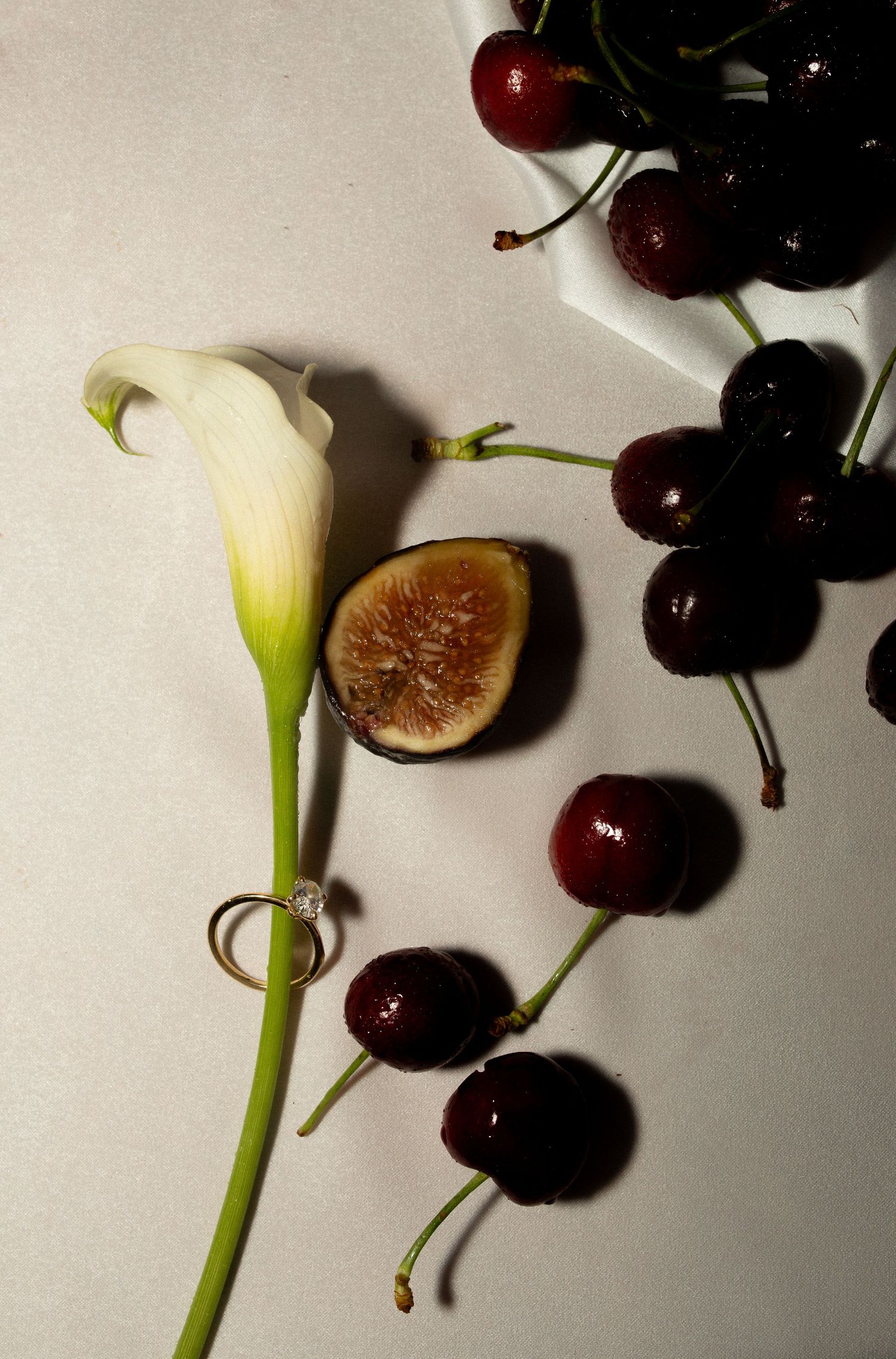 A white calla lily with a diamond ring, sliced fig, and fresh dark cherries arranged on a white surface.