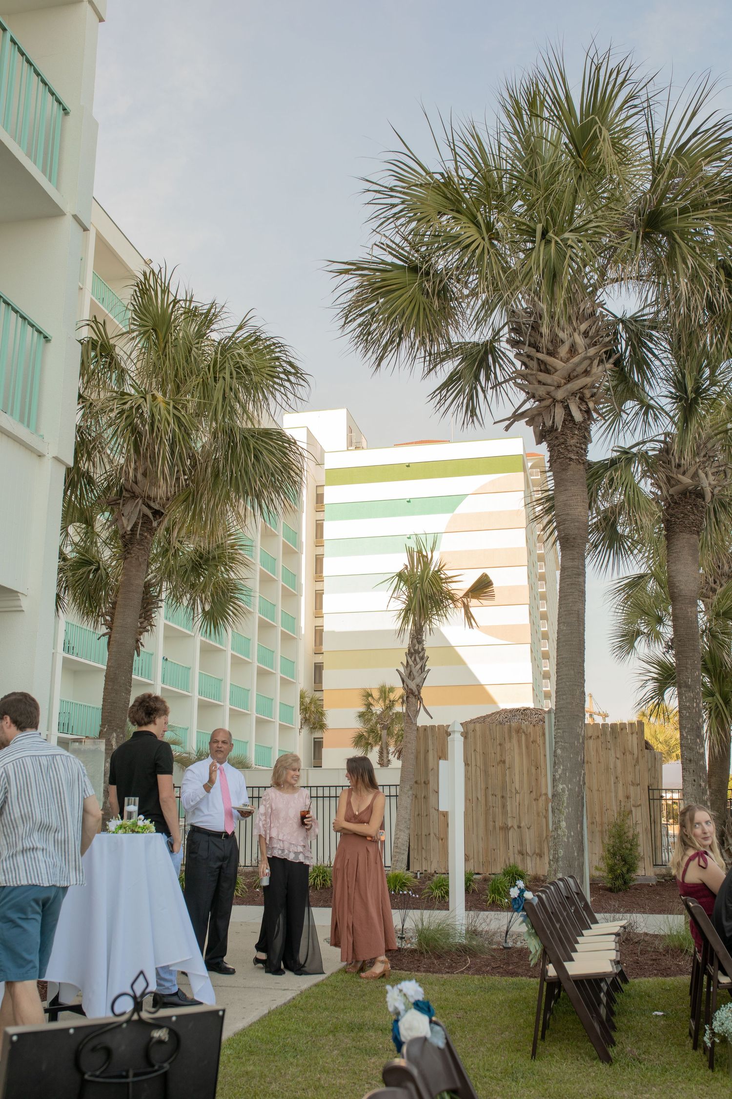 Outdoor gathering near palm trees with guests mingling beside a colorful pastel hotel building on a sunny day.