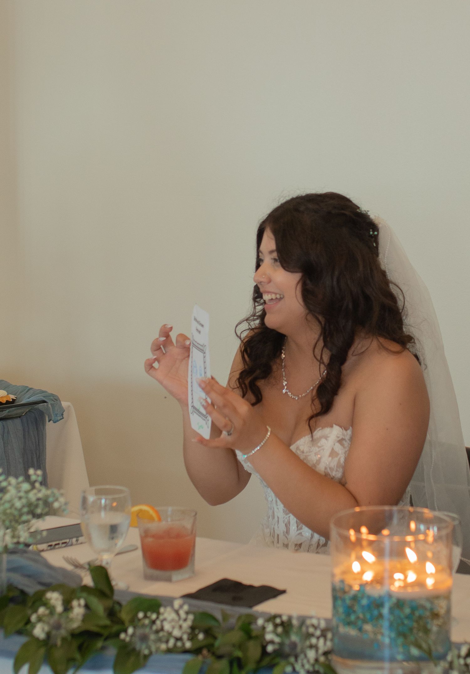 Smiling bride in strapless gown reading a card at wedding reception table with candles and floral decorations.