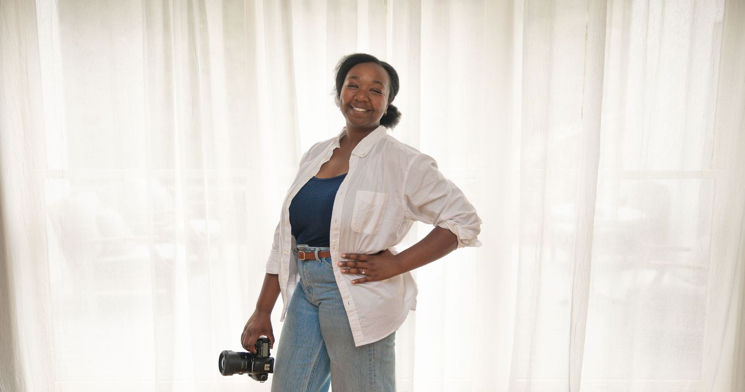 A smiling Black woman photographer holding a camera, wearing a white shirt and jeans, standing in front of sheer curtains.