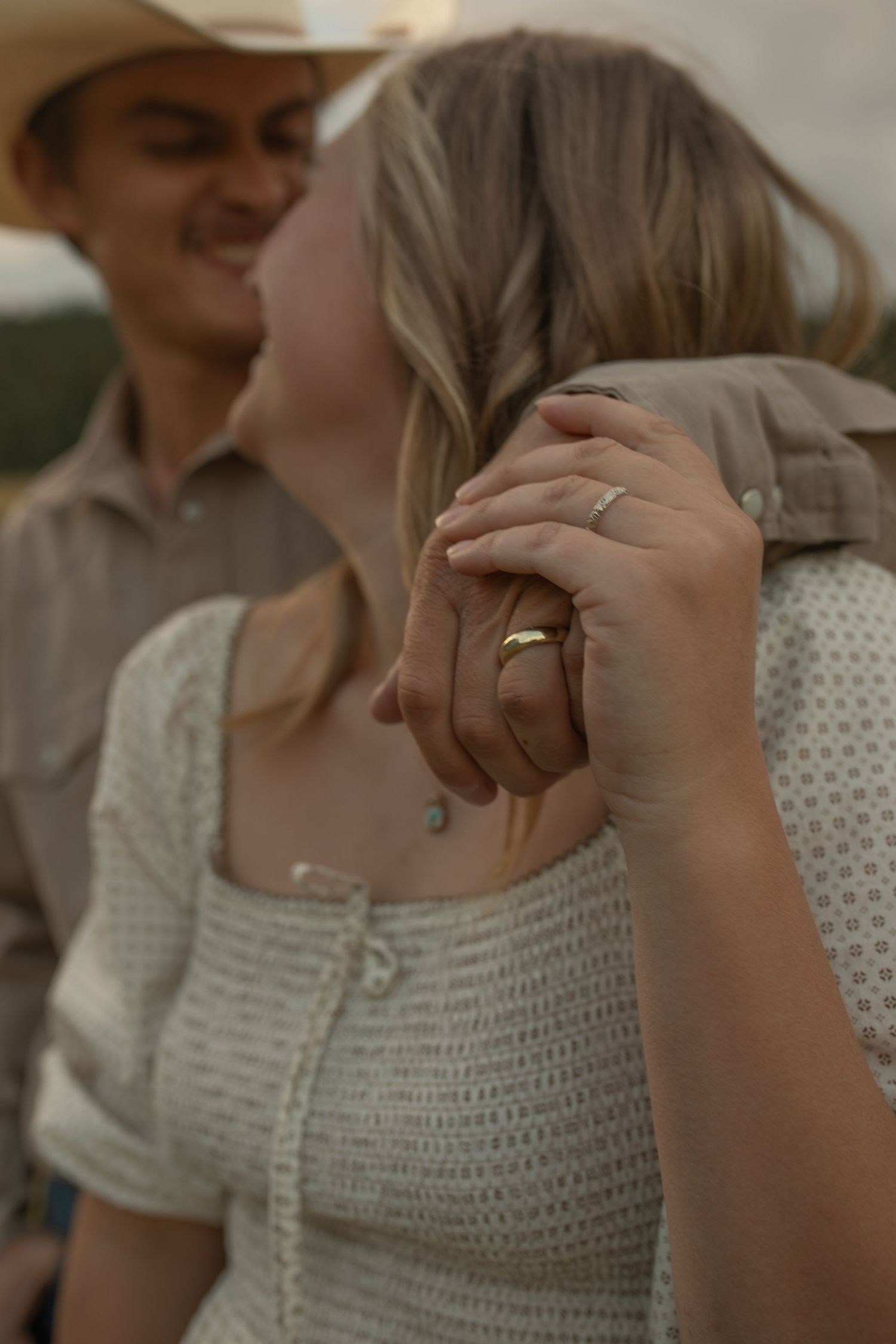 Couple holding hands closely, woman wearing gold ring in white knit top, man in cowboy hat and shirt.