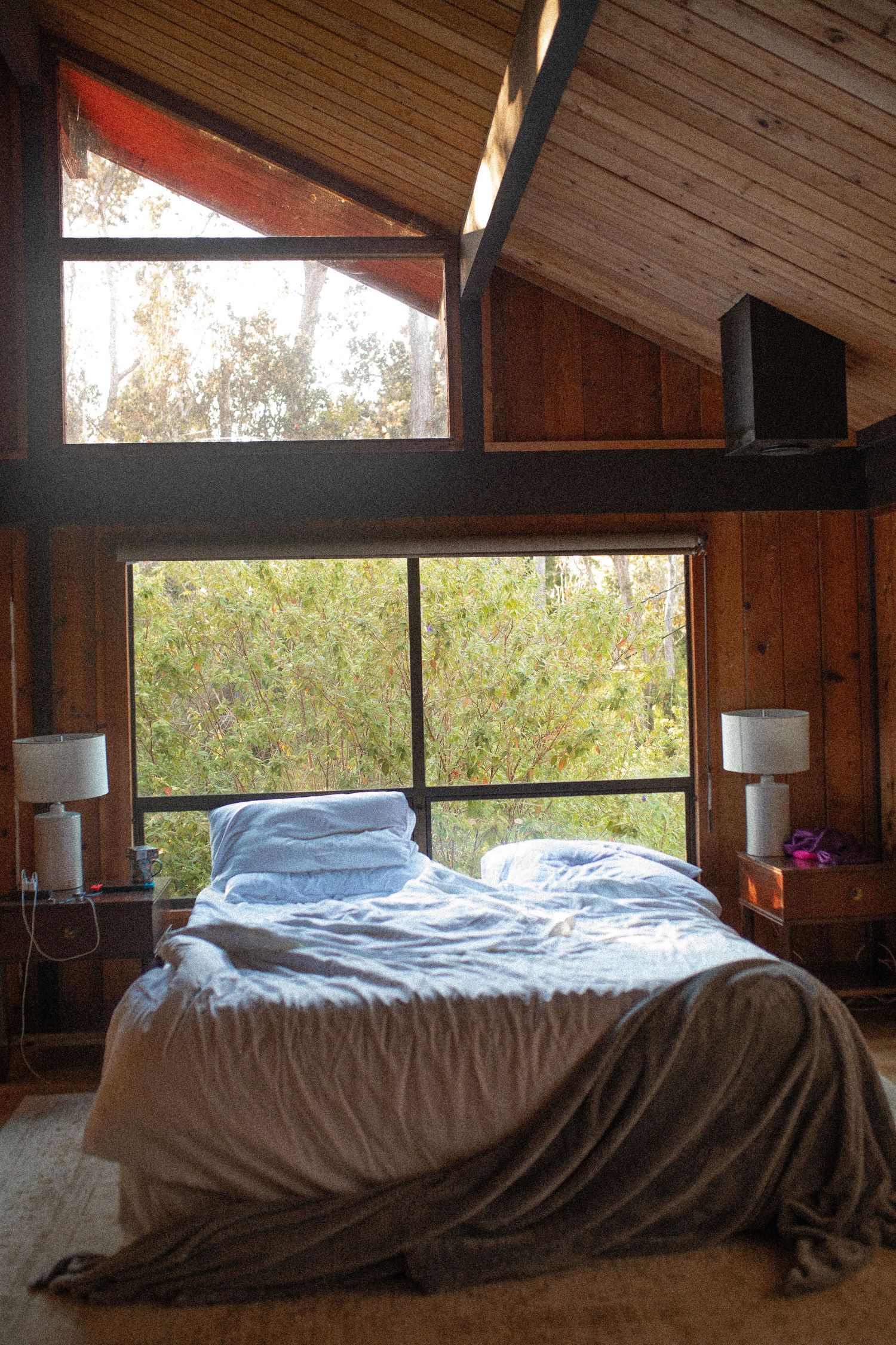Cozy bed with white linens next to large windows in rustic wooden bedroom with natural light streaming in.