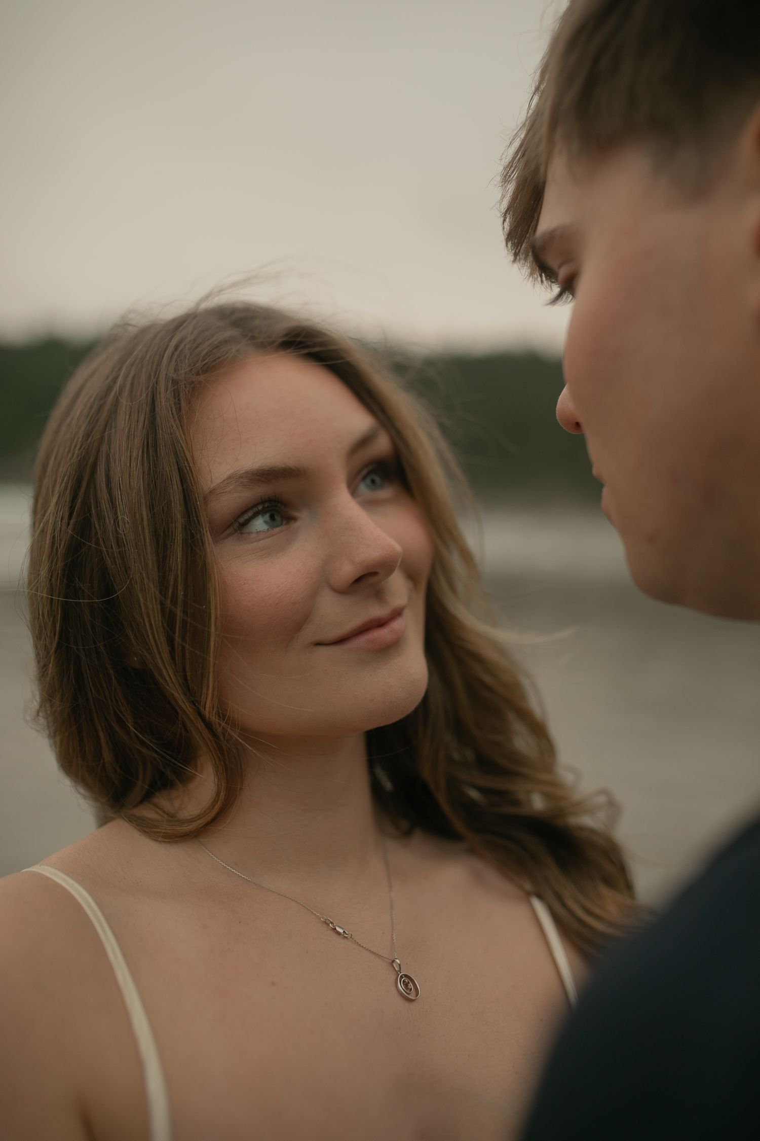 A young woman with brown hair smiles warmly at a man outdoors near water, wearing a delicate silver necklace.