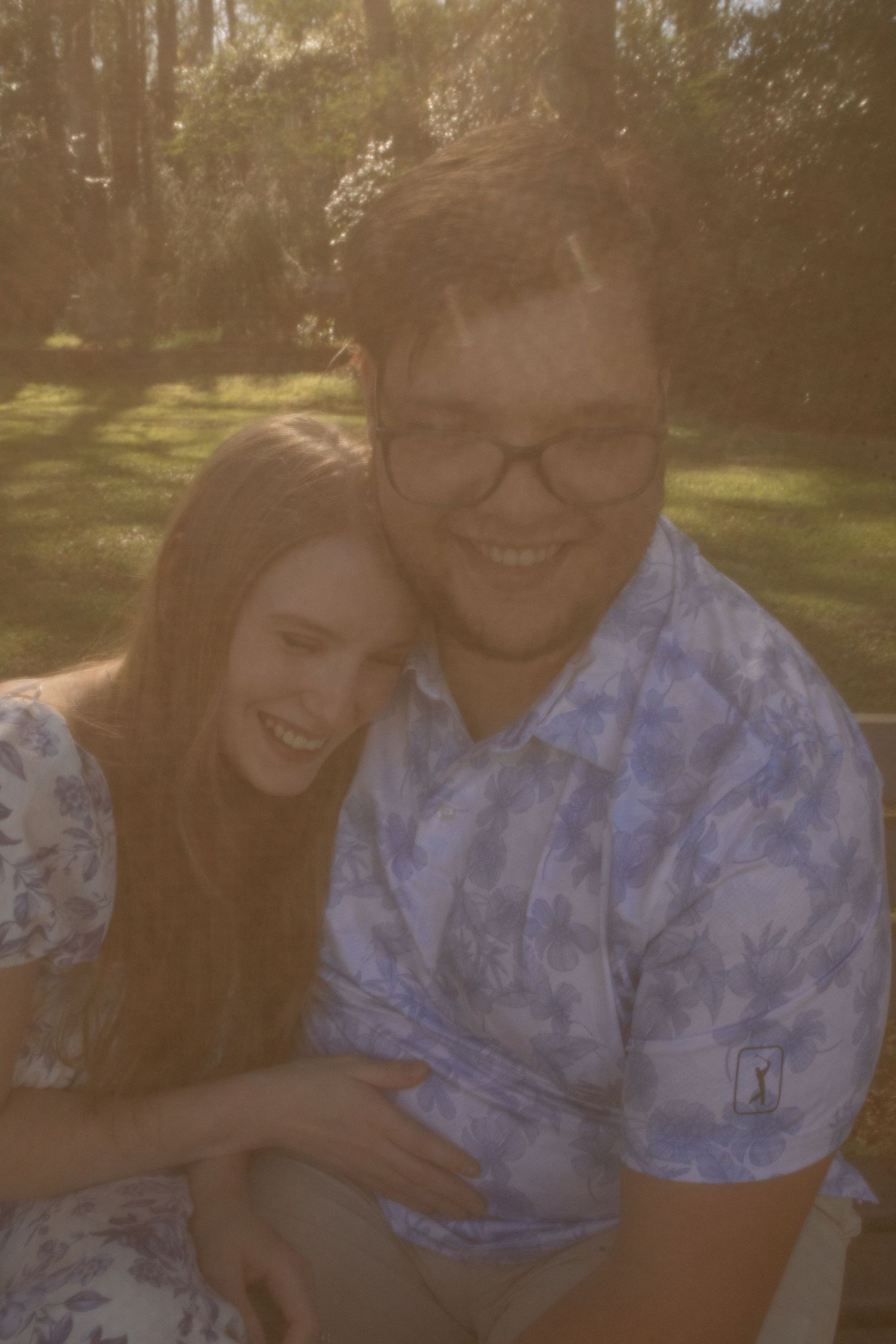 Couple smiling and embracing outdoors in golden sunlight with lush green trees in the background.