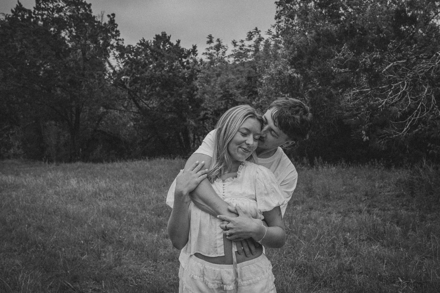 Black and white photo of a couple embracing in a grassy field surrounded by trees.