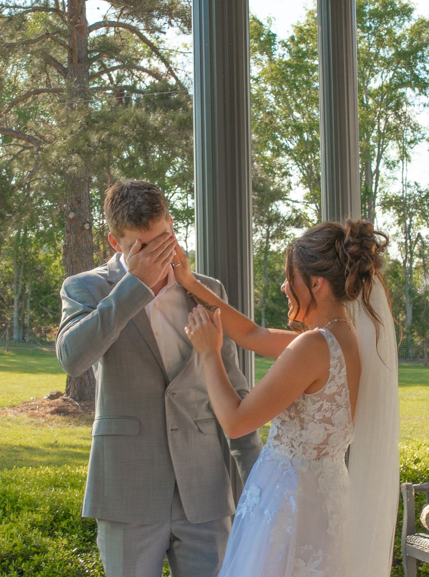 Emotional groom covering his face while bride in lace wedding dress touches his hand during first look ceremony outdoors.