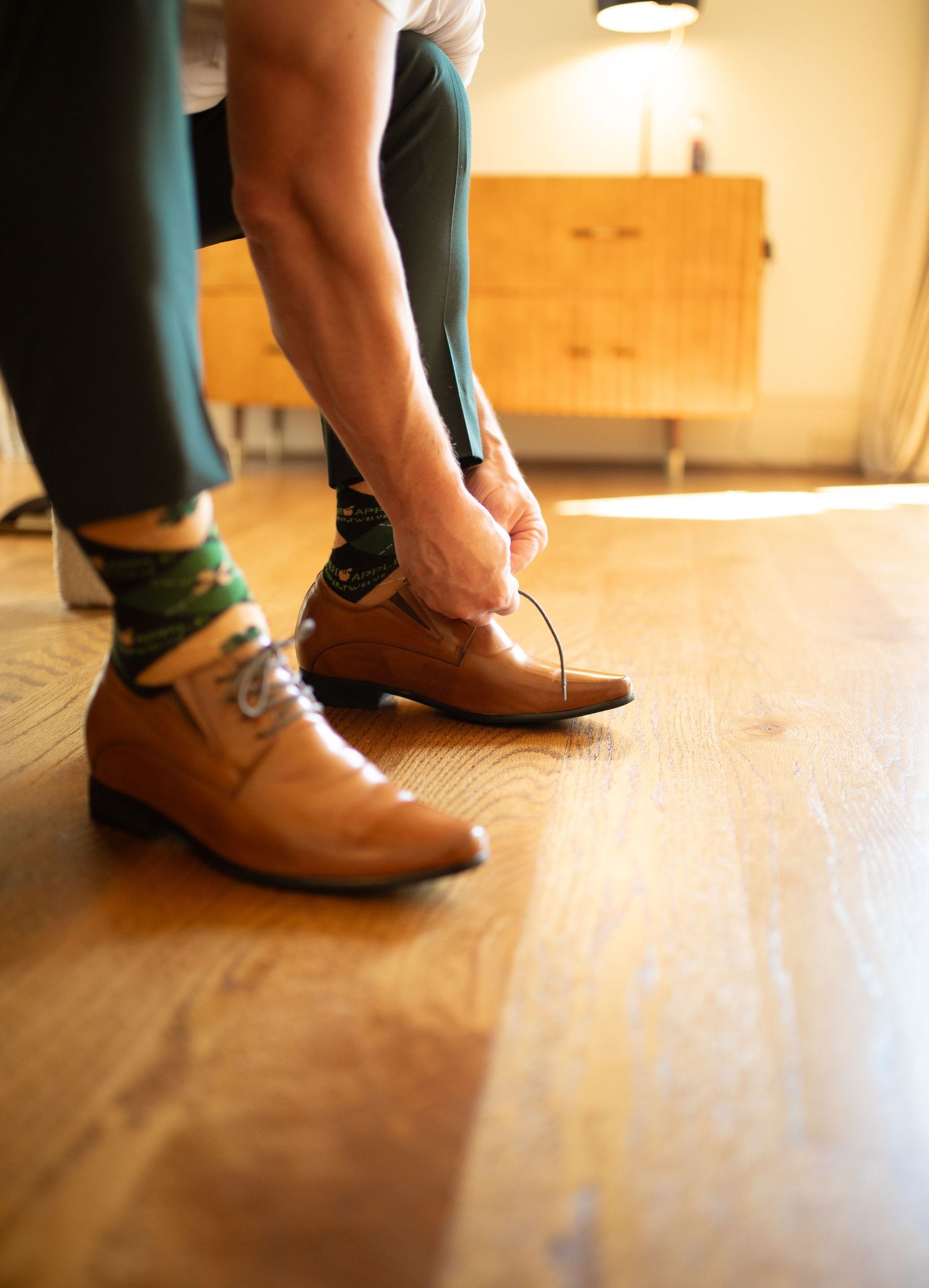 Someone tying brown leather dress shoes while wearing green socks and dark pants on a hardwood floor.