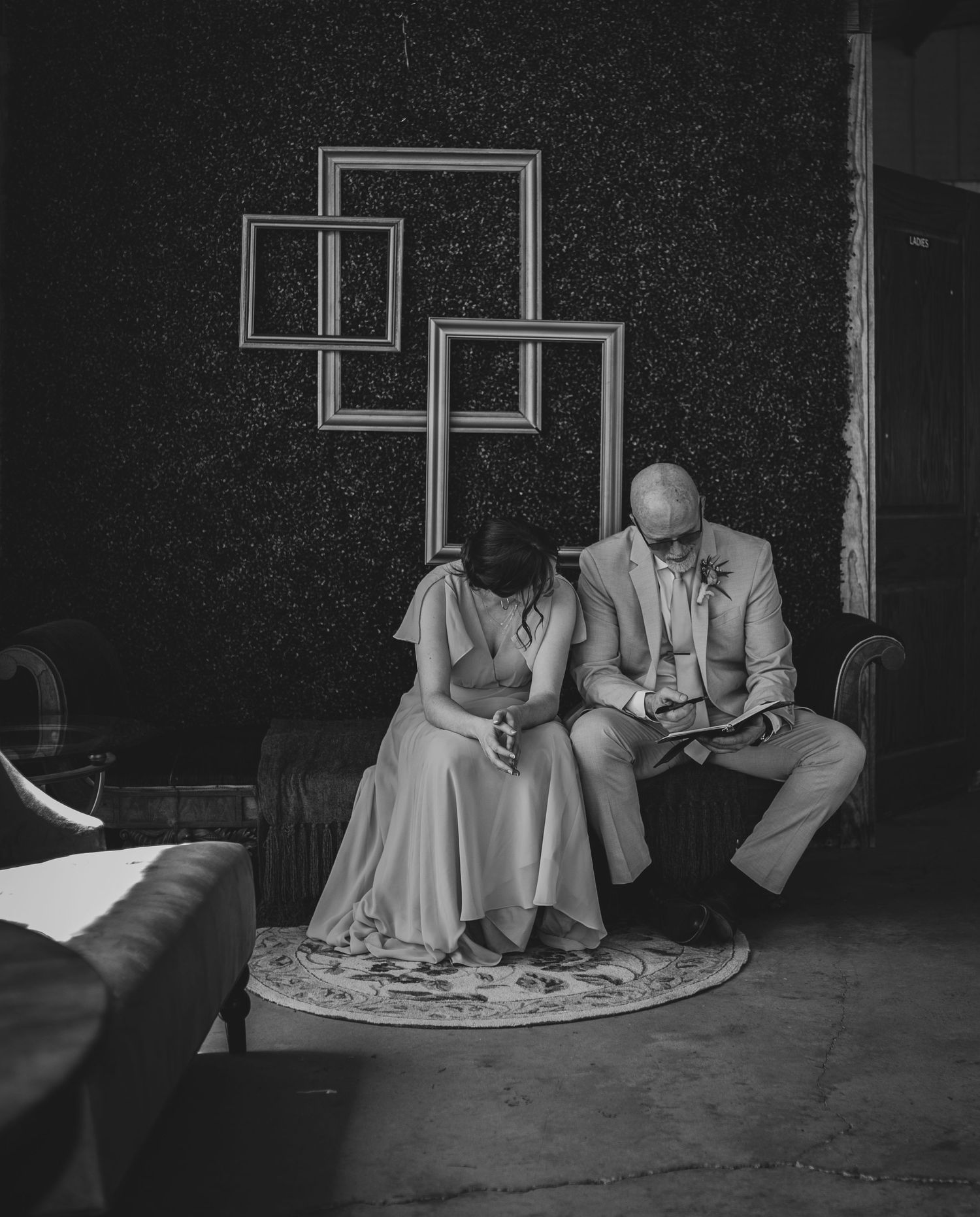 Black and white photo of a bride and groom sitting together reading vows on a floral rug at their wedding venue.