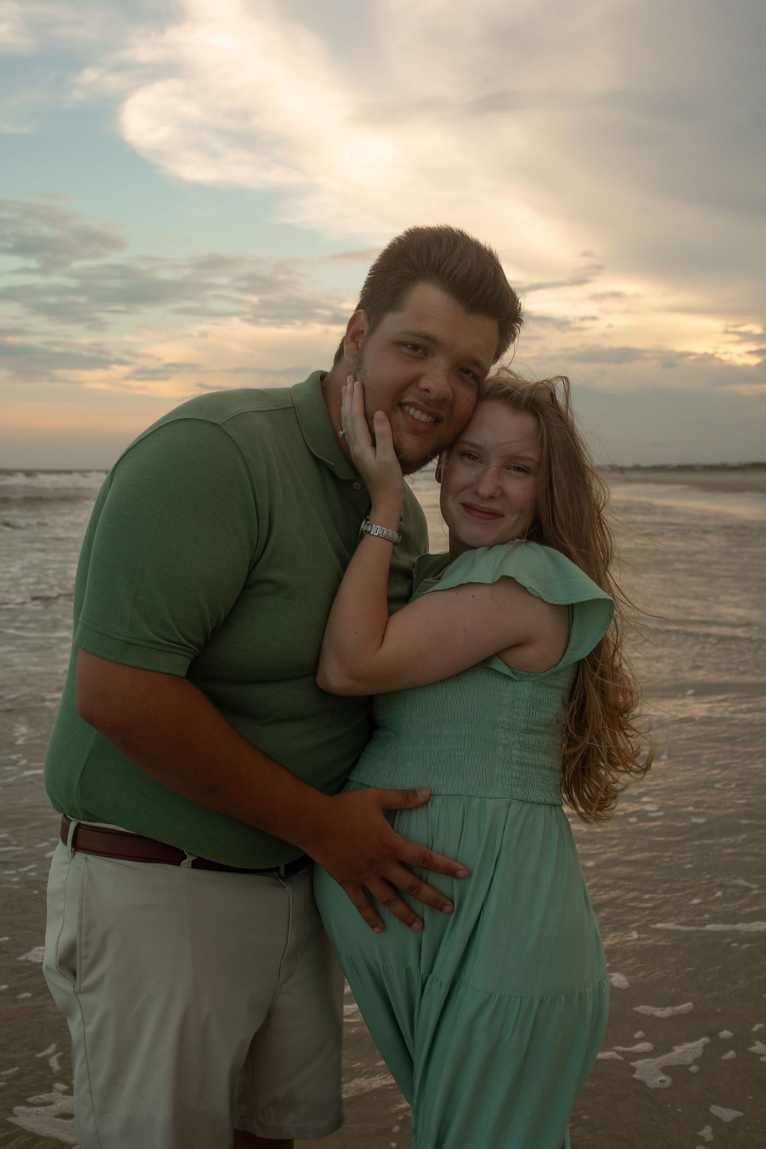 A couple embraces on the beach at sunset, wearing coordinating mint green and sage colored outfits.