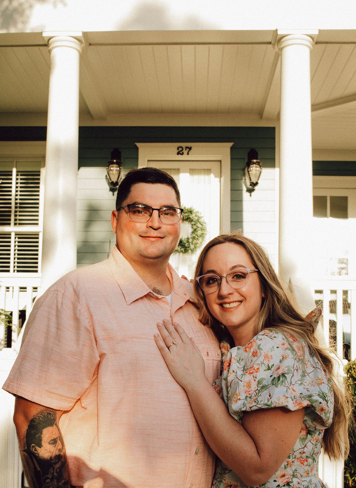 A couple wearing coordinated outfits poses on a white columned porch with house number 27.