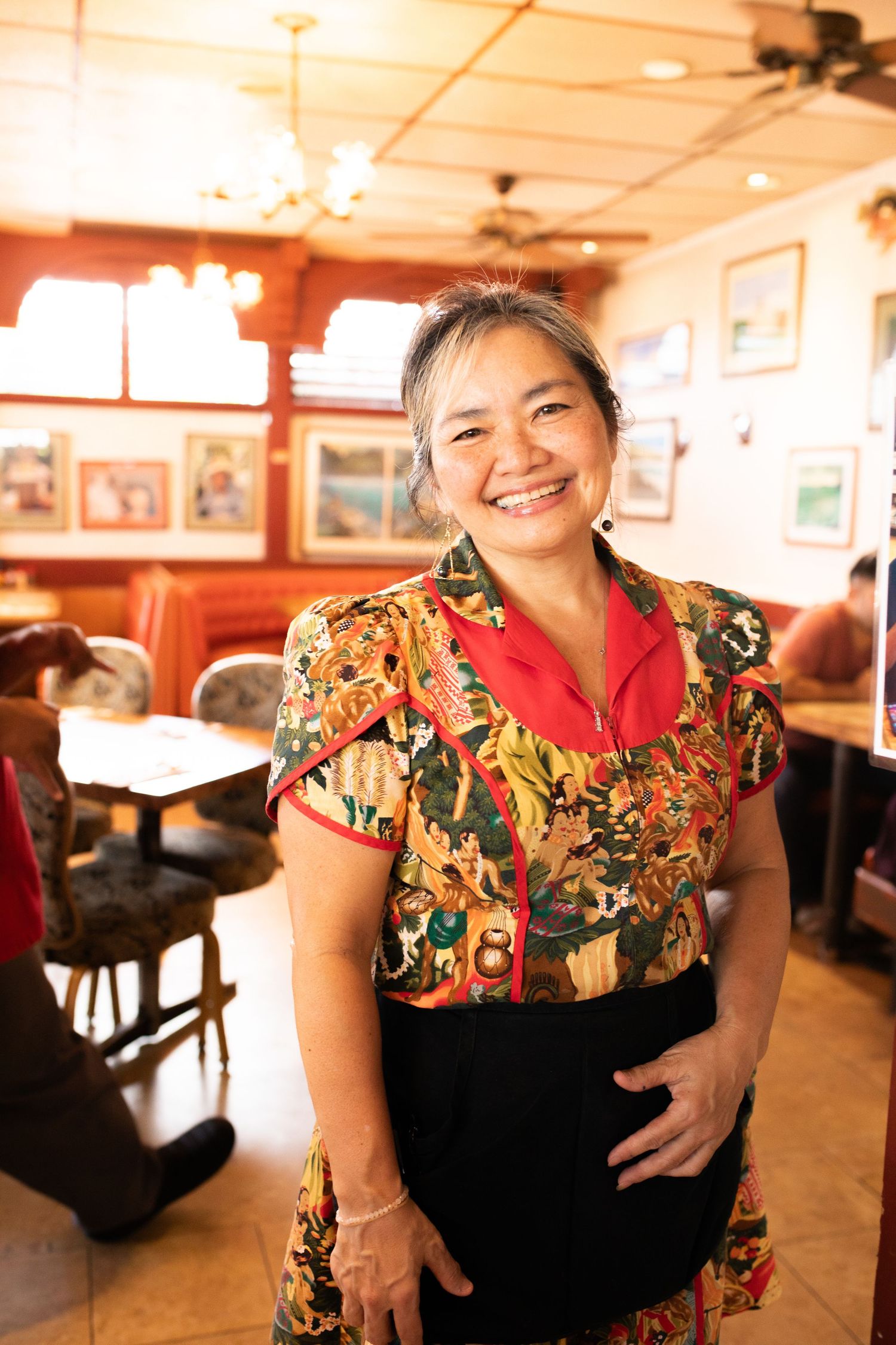 Restaurant server wearing colorful floral print dress and red collar smiles warmly in casual dining room setting.