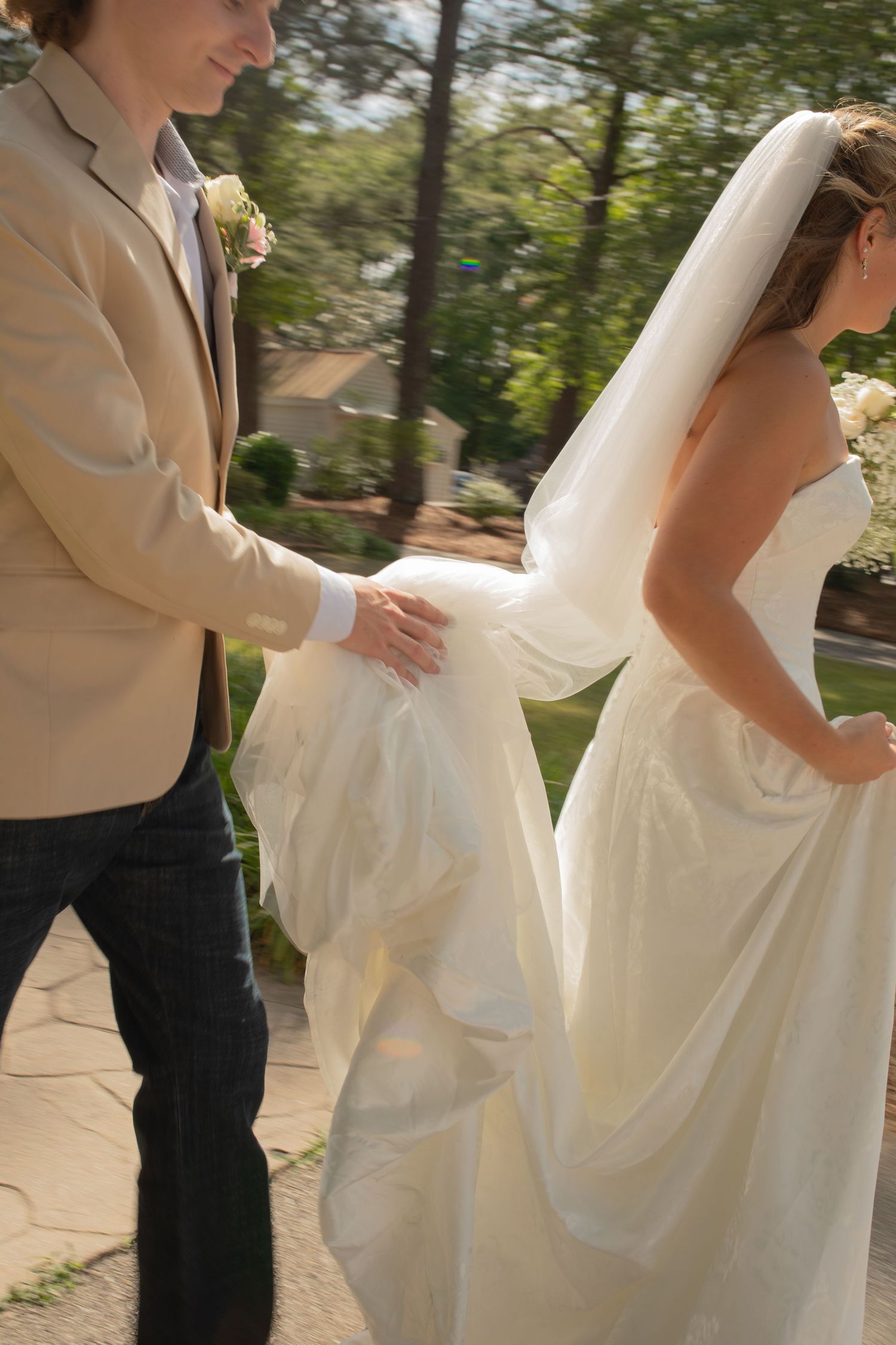 Groom in tan jacket helps bride carry her white wedding gown train outdoors on a sunny day.